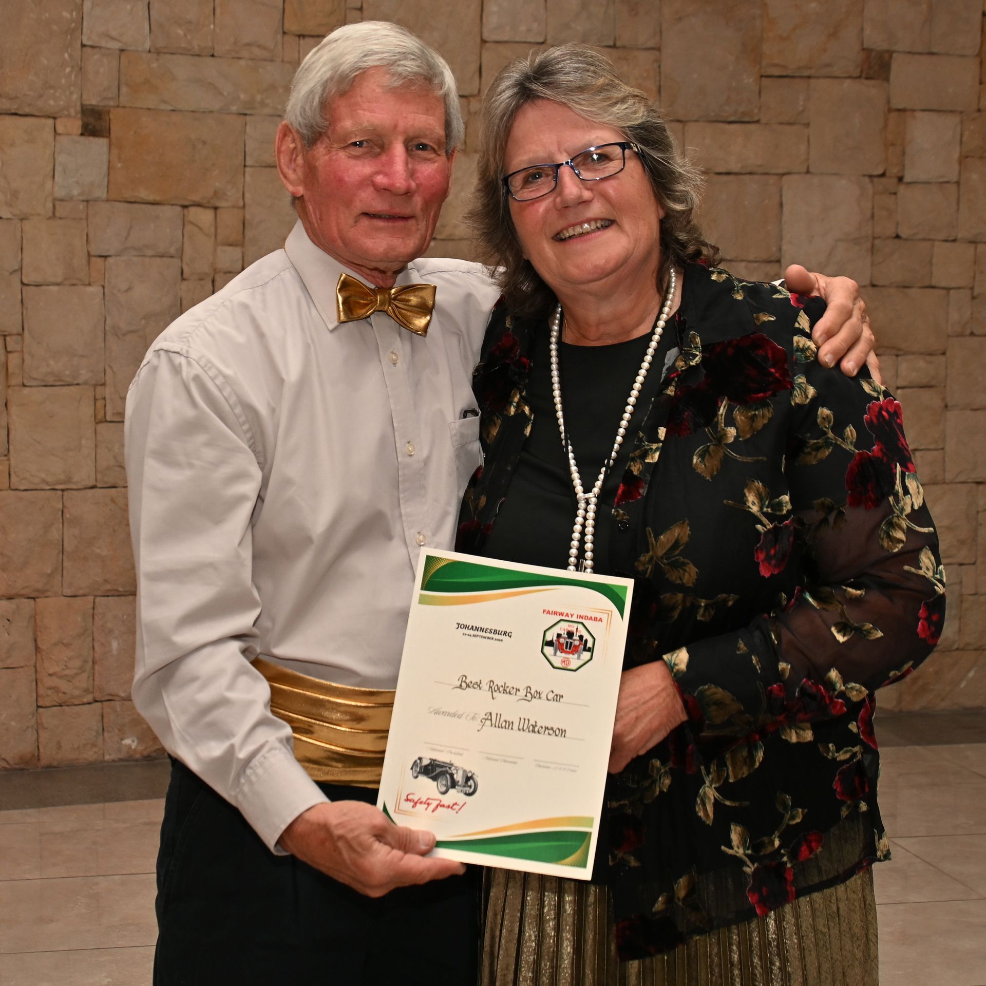 Two people smiling, holding a certificate, standing before a stone wall.