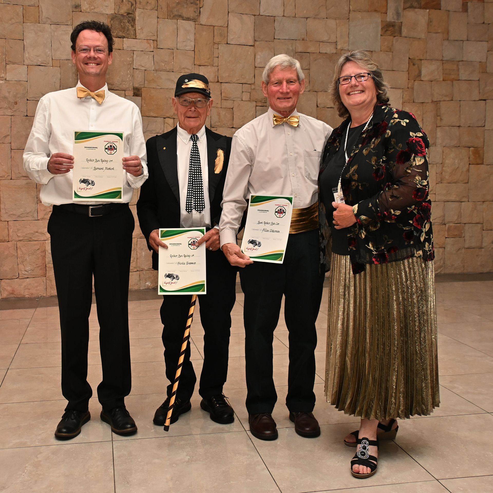 Four people pose indoors in formal attire, three holding certificates in front of a stone wall.