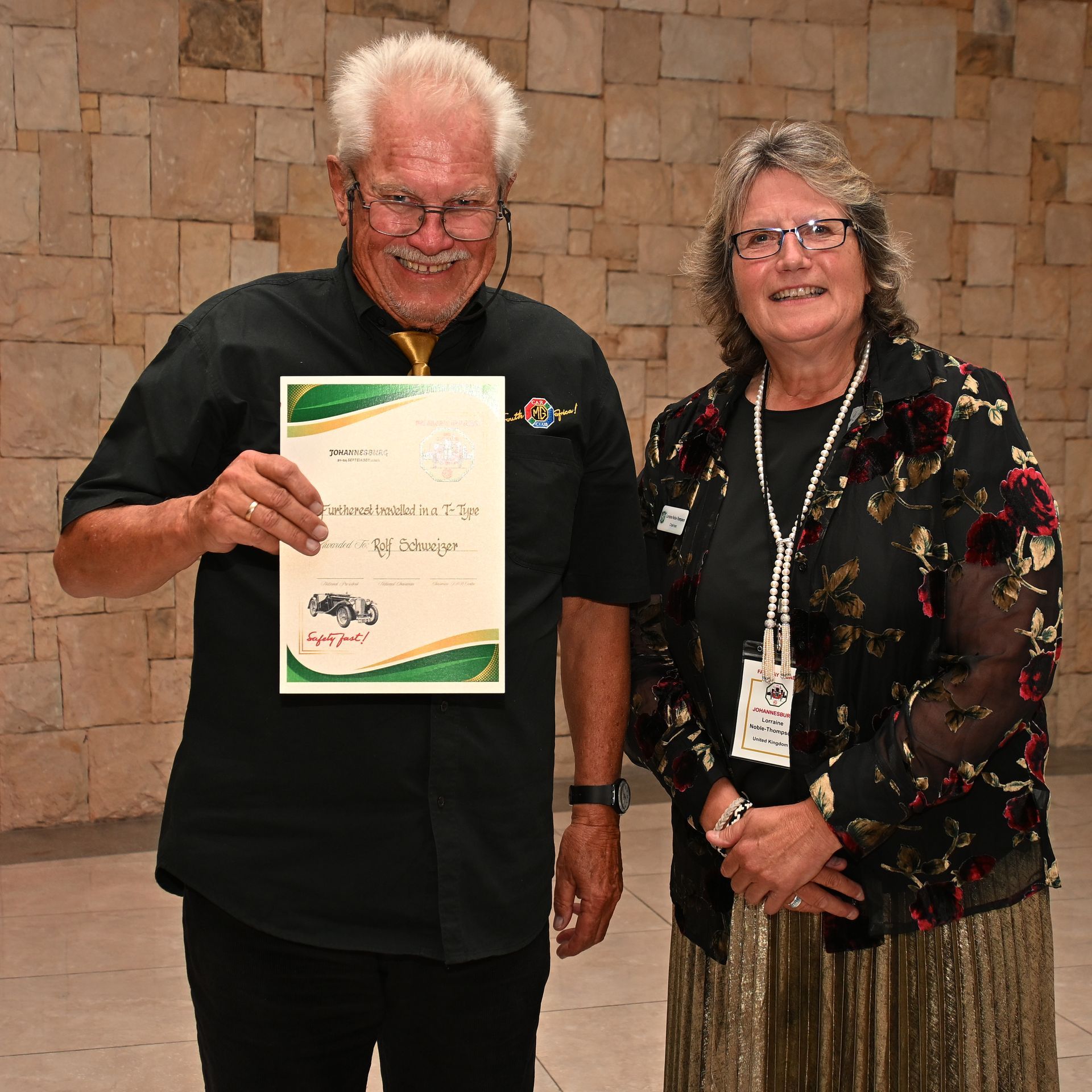 Two people standing by a stone wall, one holding a certificate and both smiling.