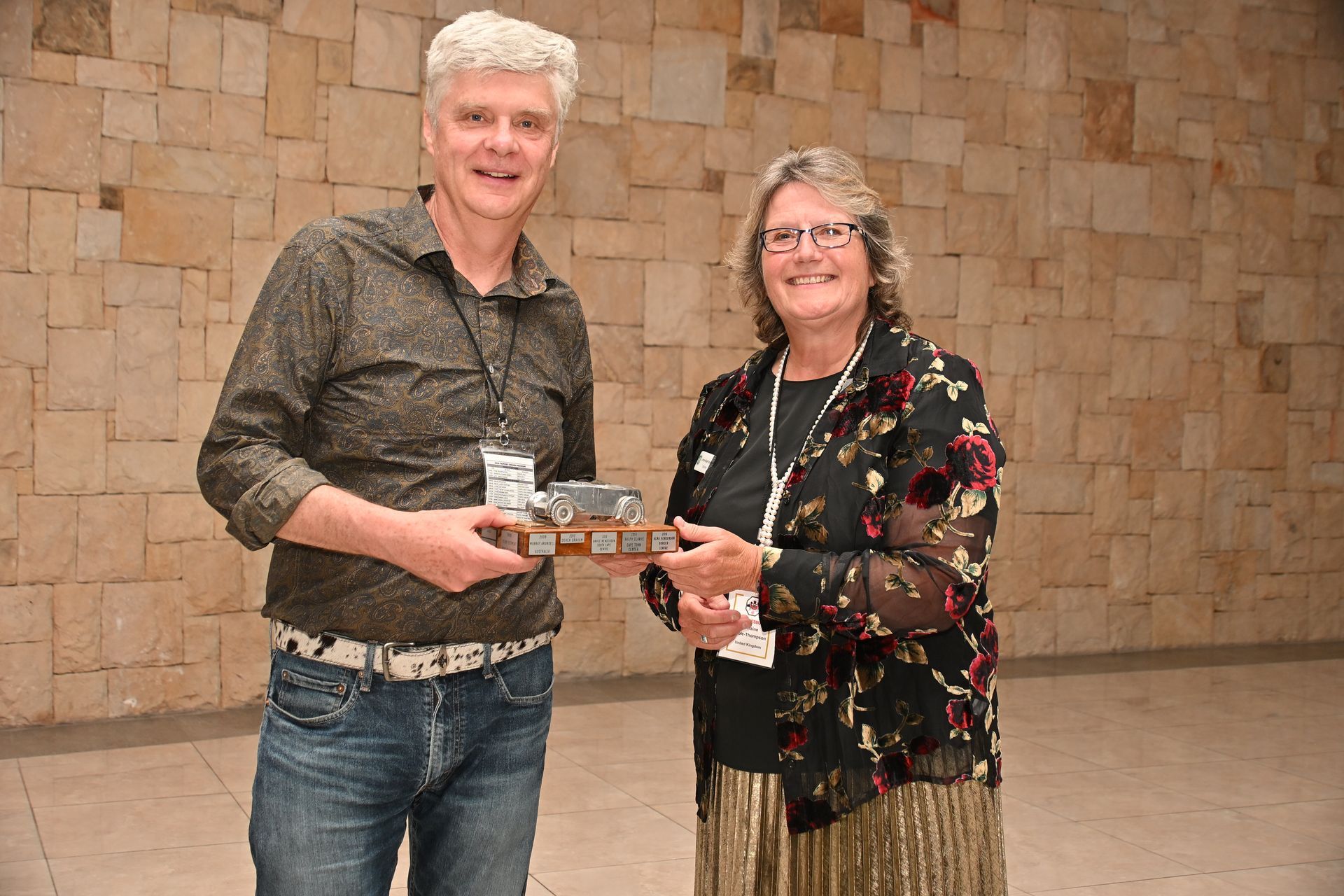Two people holding a small award plaque, standing in front of a stone wall and smiling