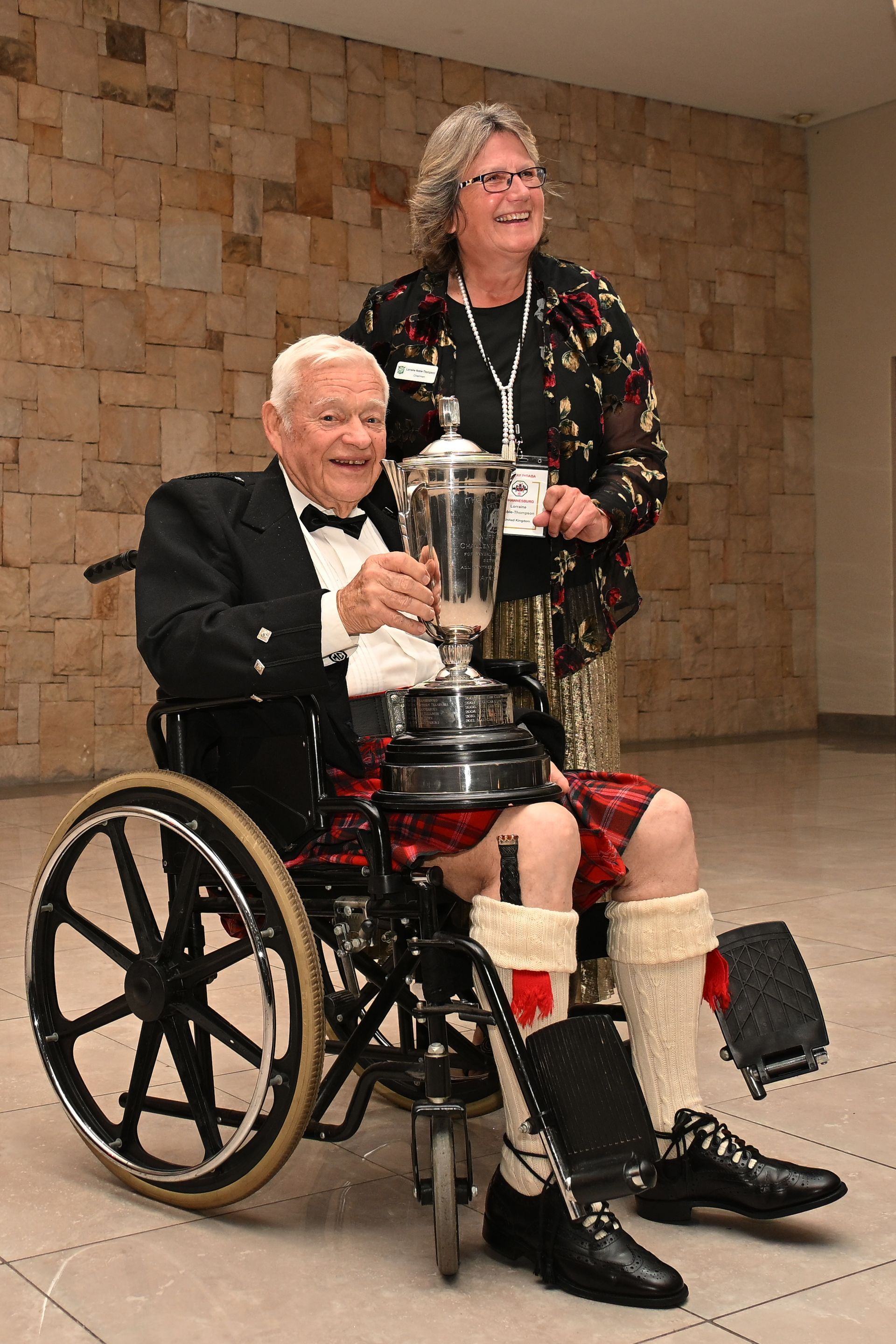 Man in a wheelchair holding a trophy, with a woman standing behind him in a hallway