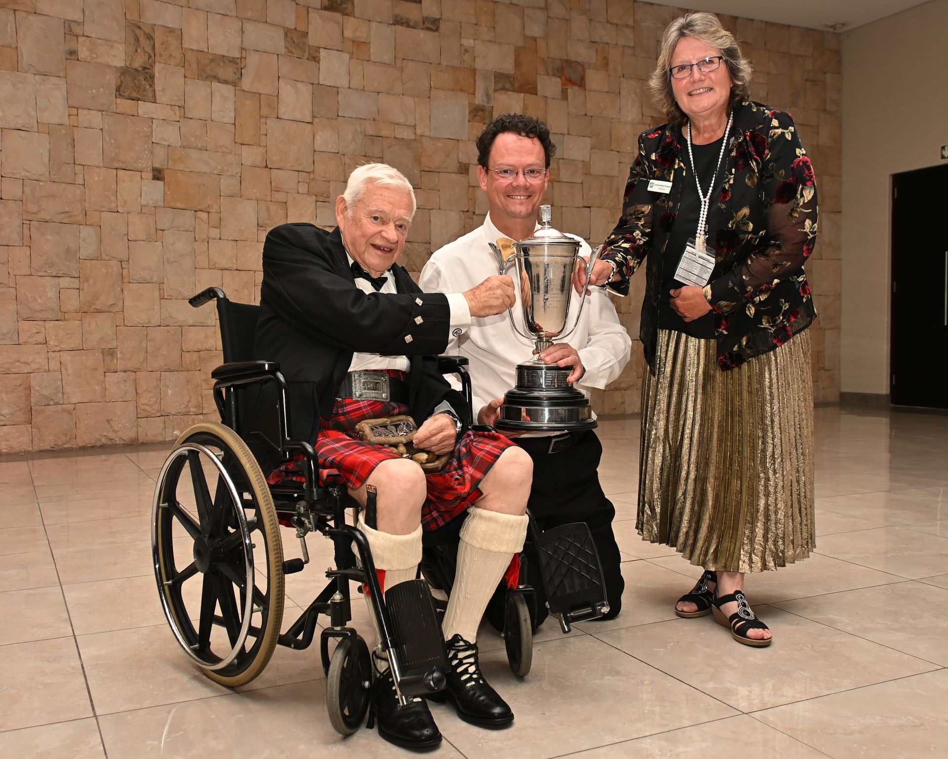 Three people pose indoors with a trophy; one sits in a wheelchair.