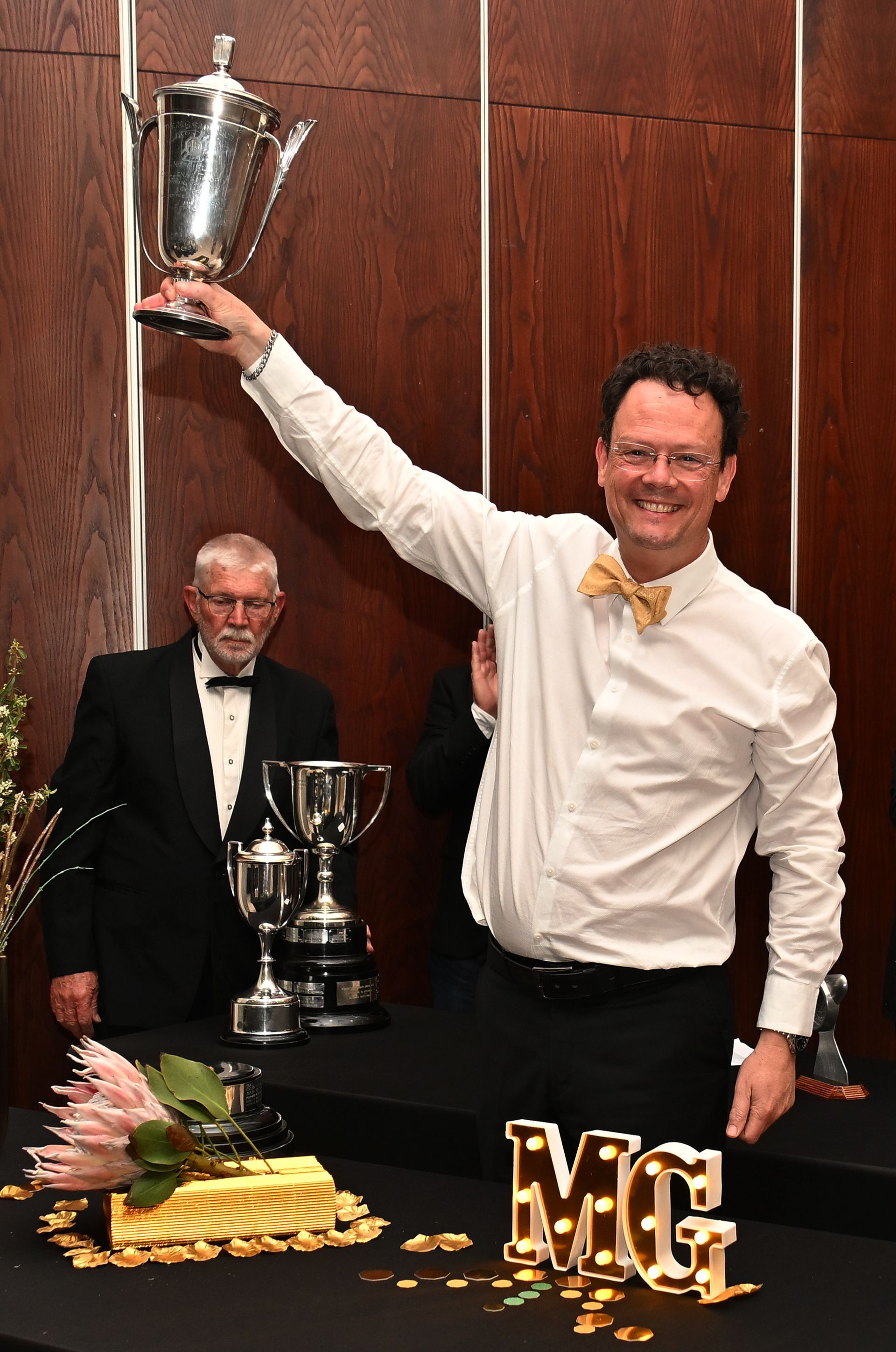 Man in white shirt holding up a silver trophy at a formal awards event, with another man and trophies behind him.