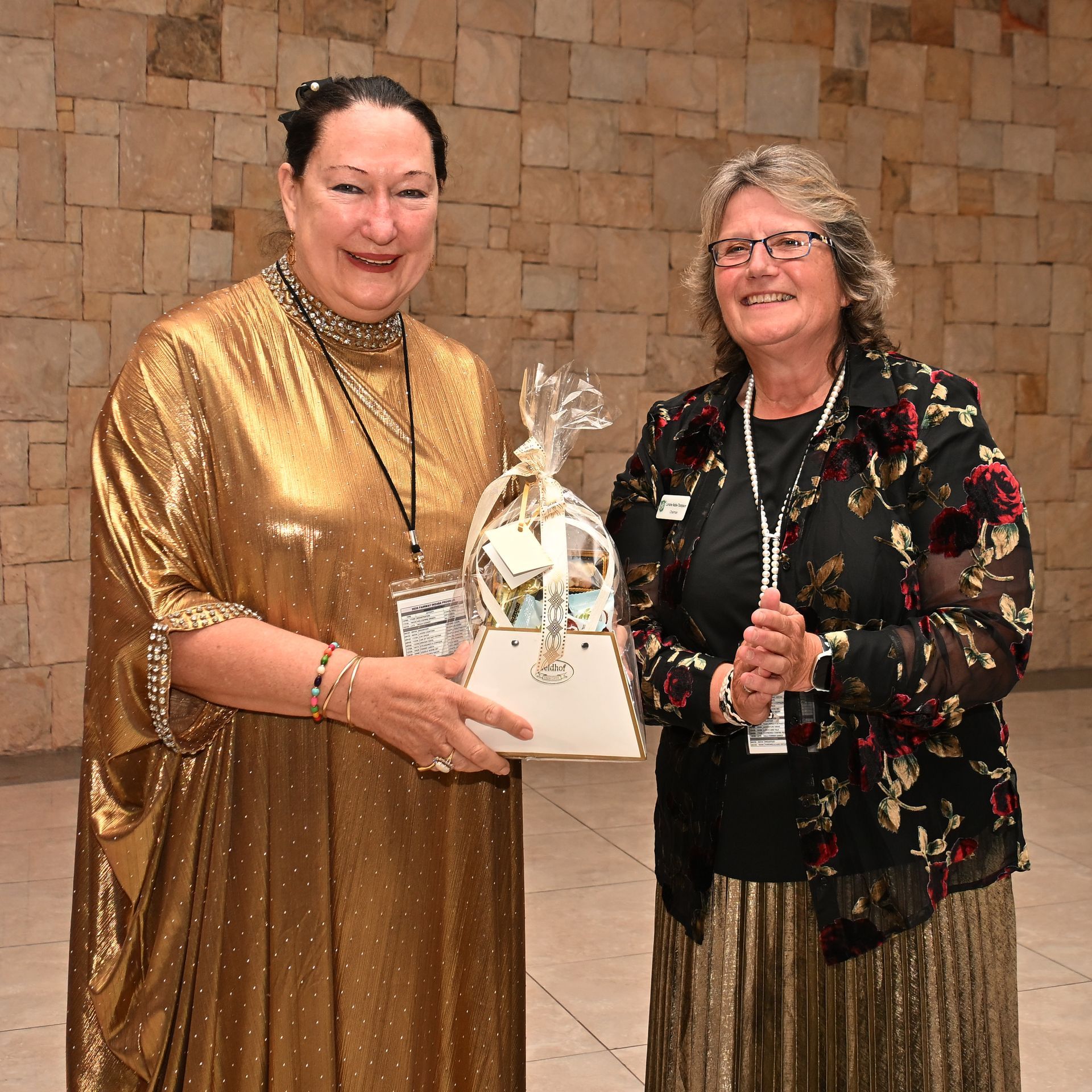 Two women holding a wrapped gift basket, smiling in a brick hall.