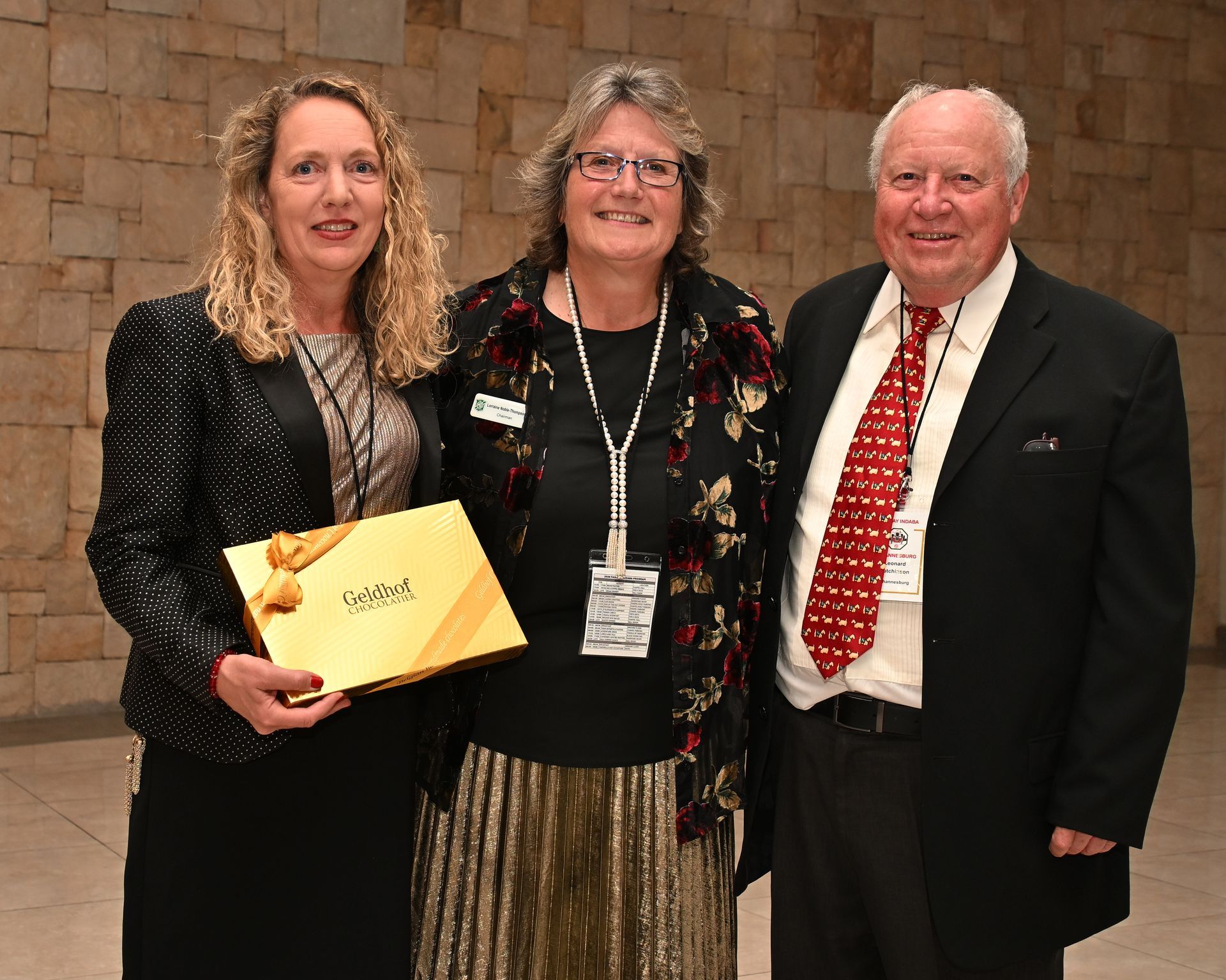 Three people posing indoors; one holds a yellow envelope, all wearing formal attire.