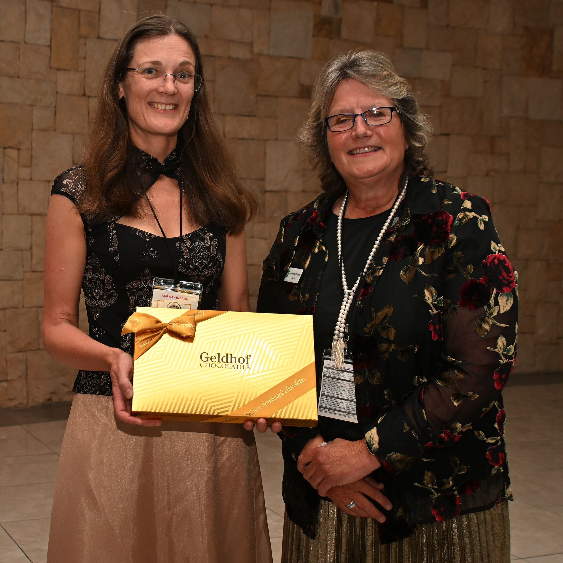 Two women smiling and holding a gold gift box at an indoor event