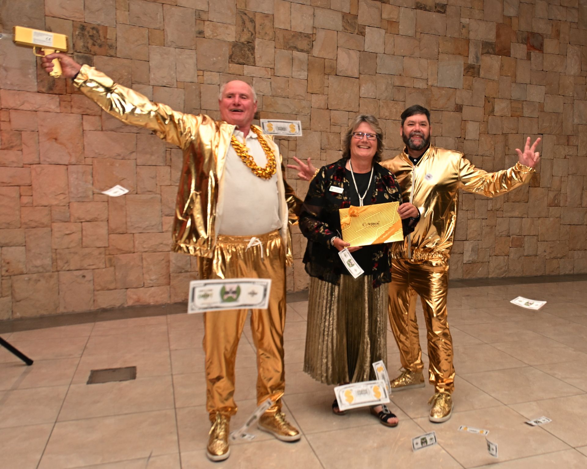 Three people in gold outfits pose holding awards in a stone-walled lobby.