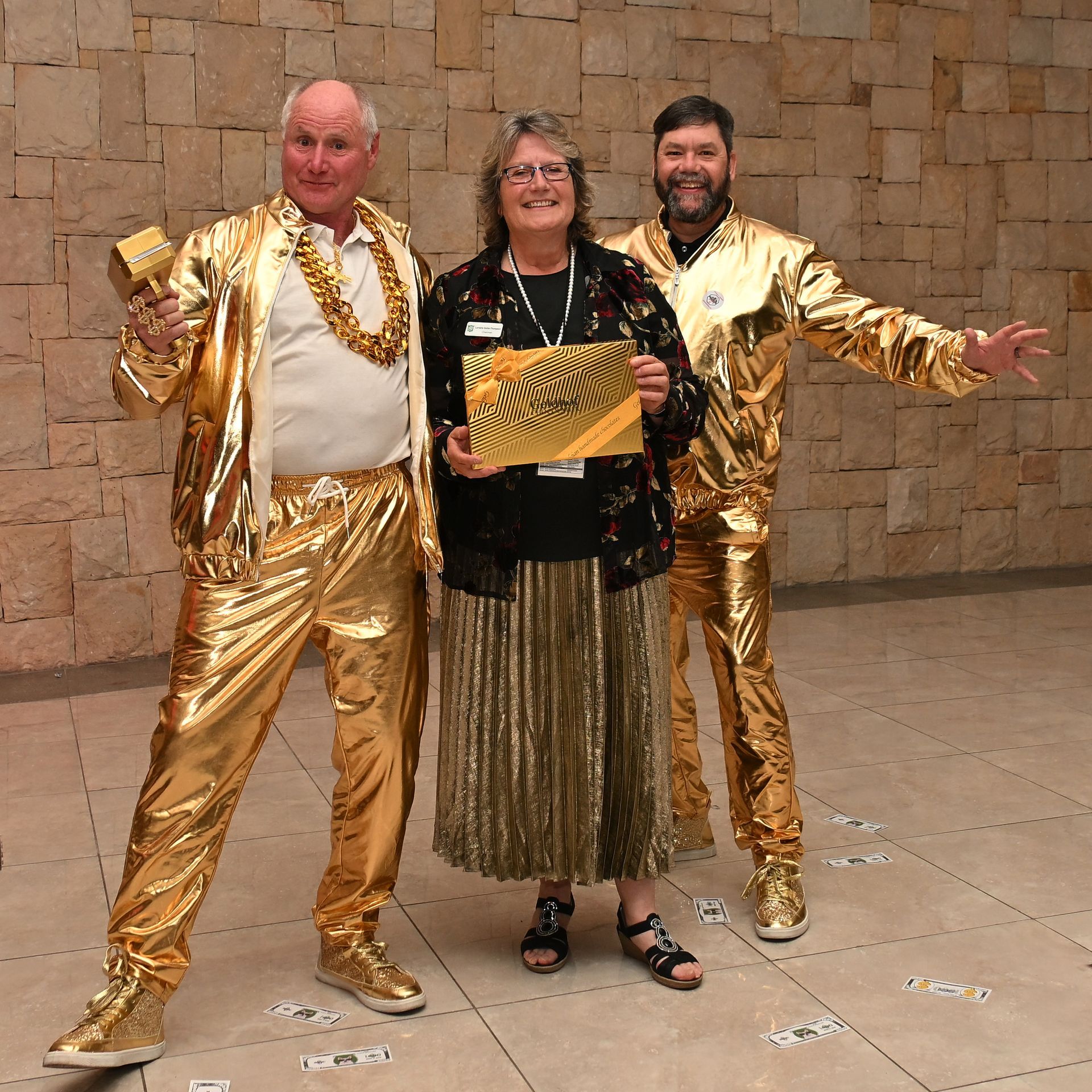 Three people in gold outfits posing indoors, holding a gold award plaque against a stone wall.