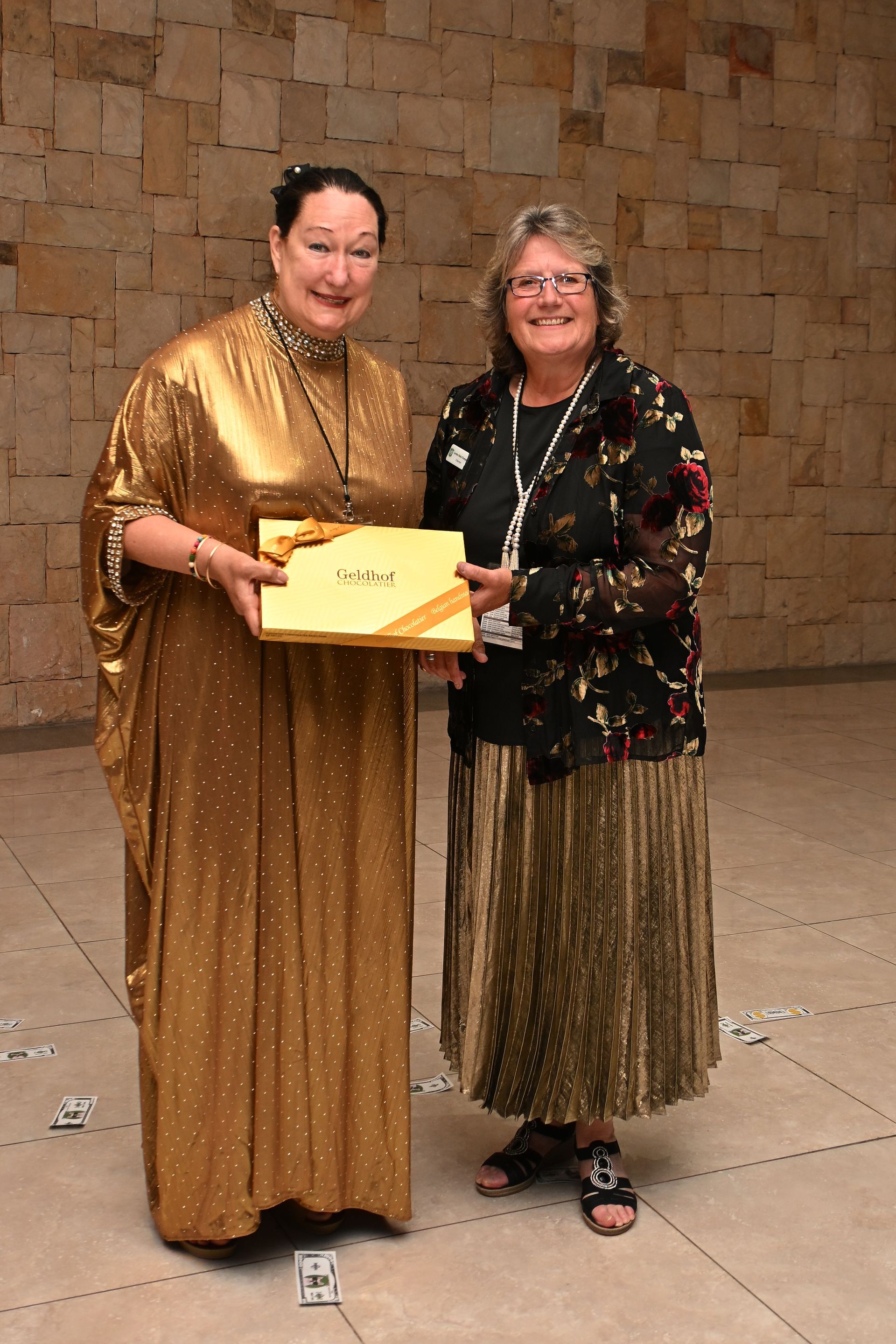 Two women pose indoors, holding a gold award plaque, one in a gold dress and one in a floral jacket.