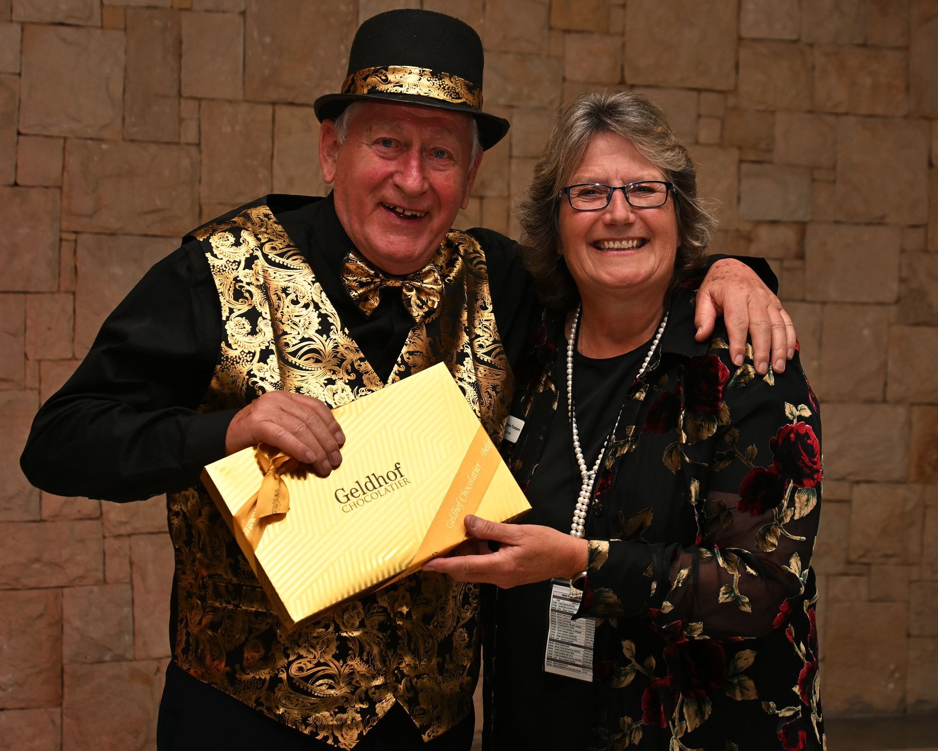 Two smiling people posing indoors, one holding a yellow envelope and wearing a black-and-gold vest.
