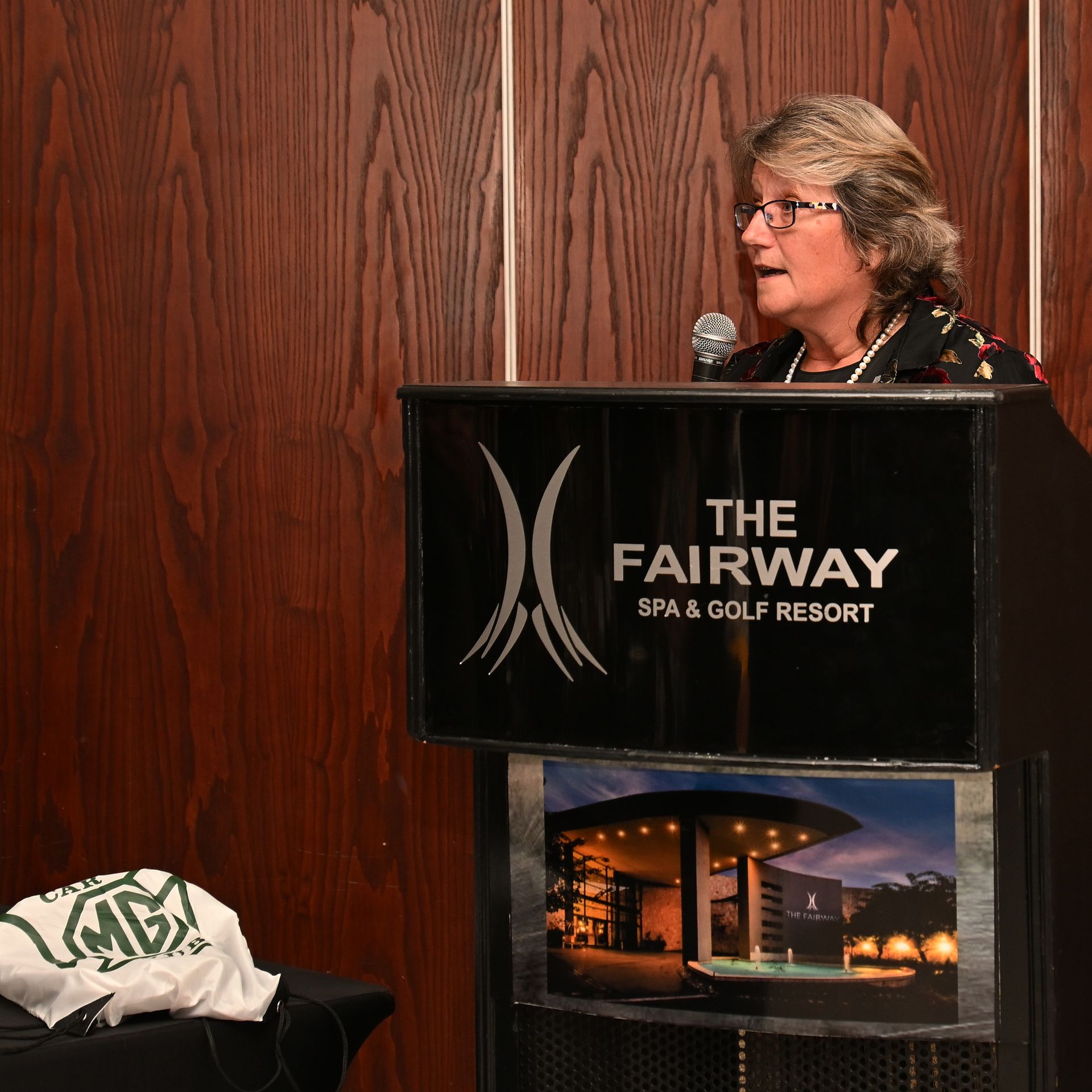 Woman speaking at a podium with The Fairway Spa & Golf Resort sign in a wood-paneled room