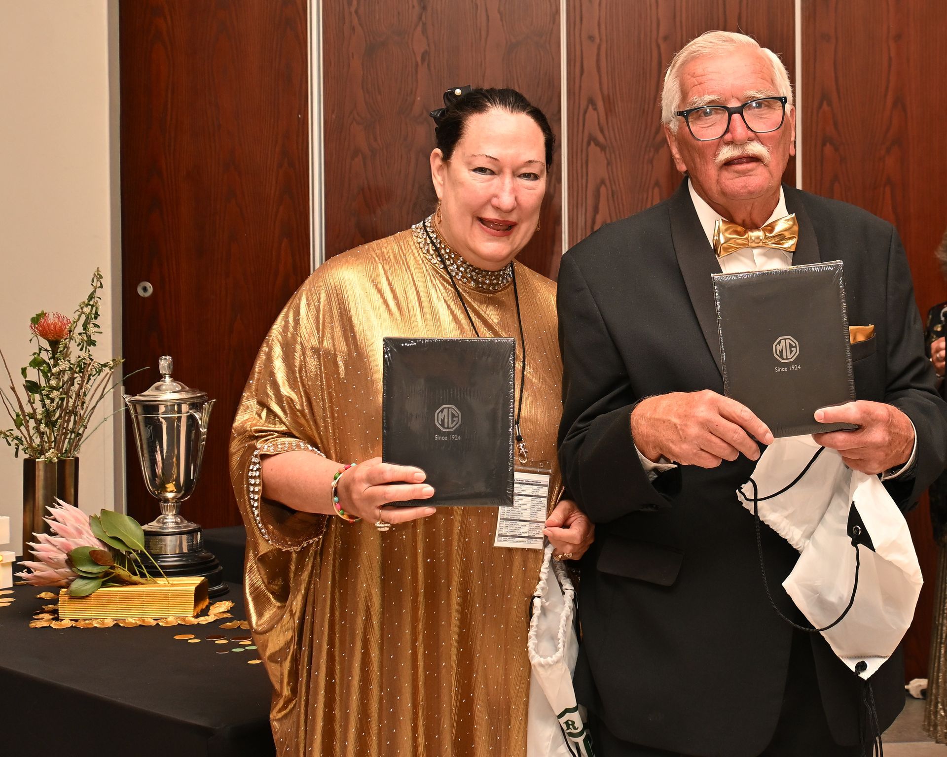 Two people in formal wear hold framed awards at a ceremony table with a trophy