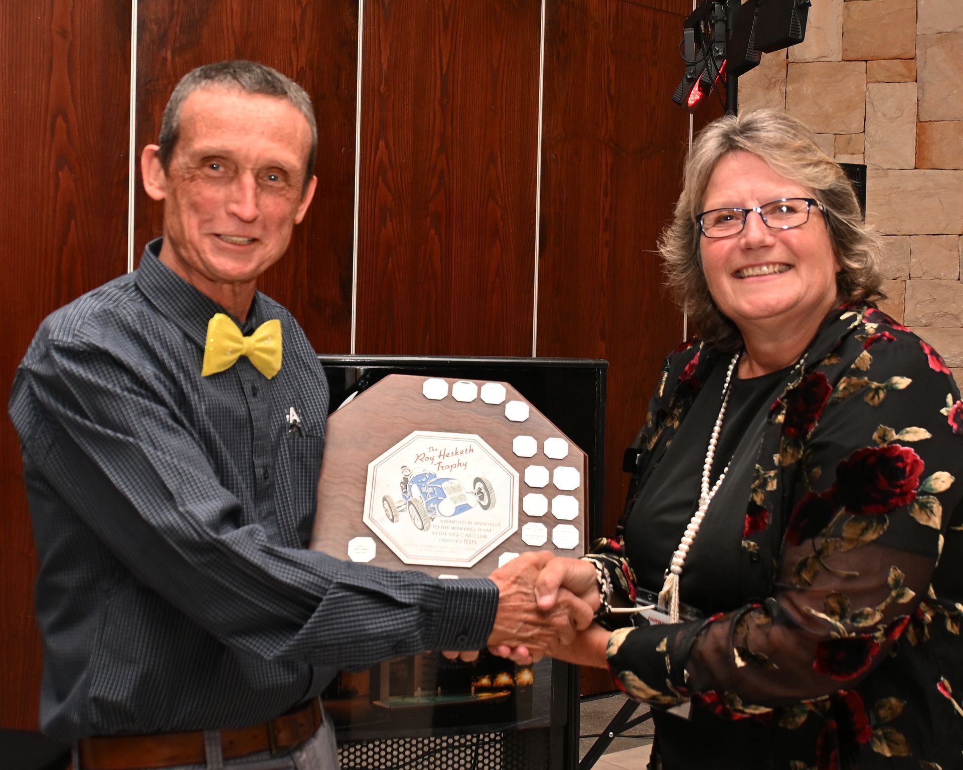 Two people shake hands while holding an award plaque in front of a wooden wall.