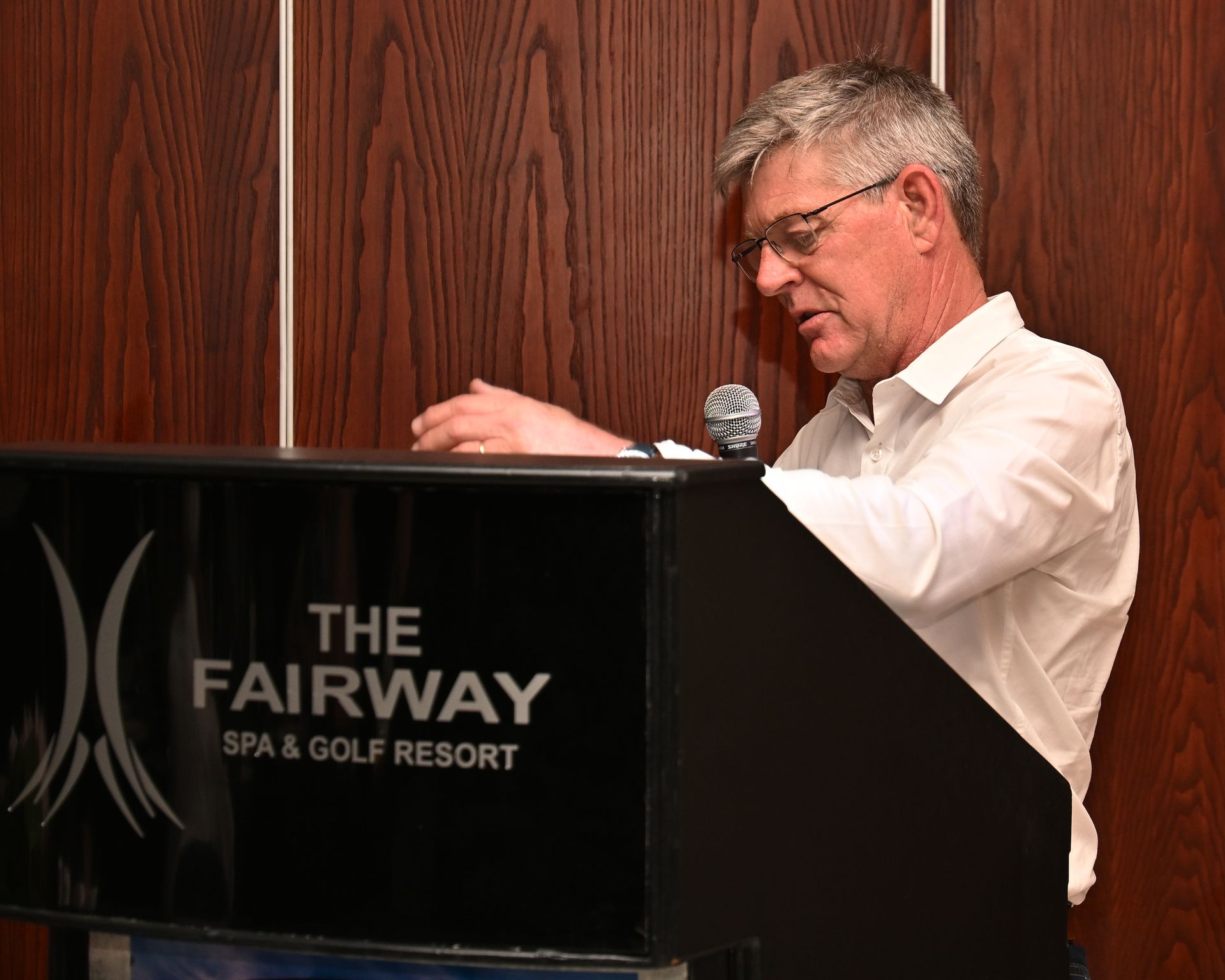 Man speaking at a podium labeled “The Fairway Spa & Golf Resort” against a wood-paneled wall