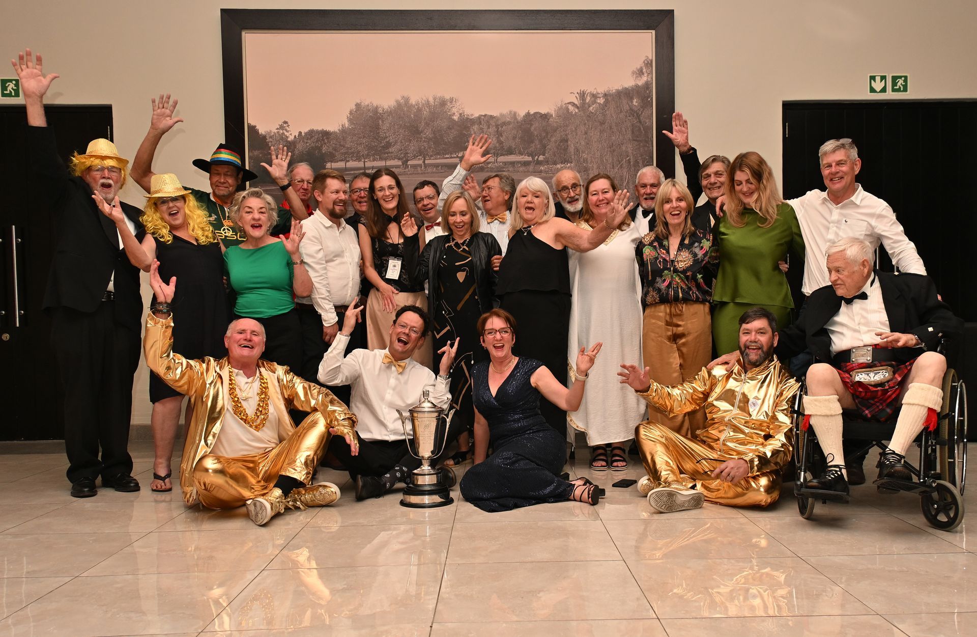 Group of people posing and cheering indoors, with several in gold costumes and two seated in wheelchairs.