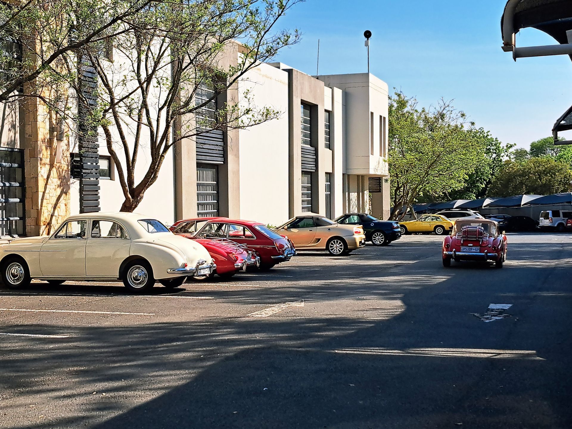 Parking lot with several parked cars beside a beige building and trees on a sunny day