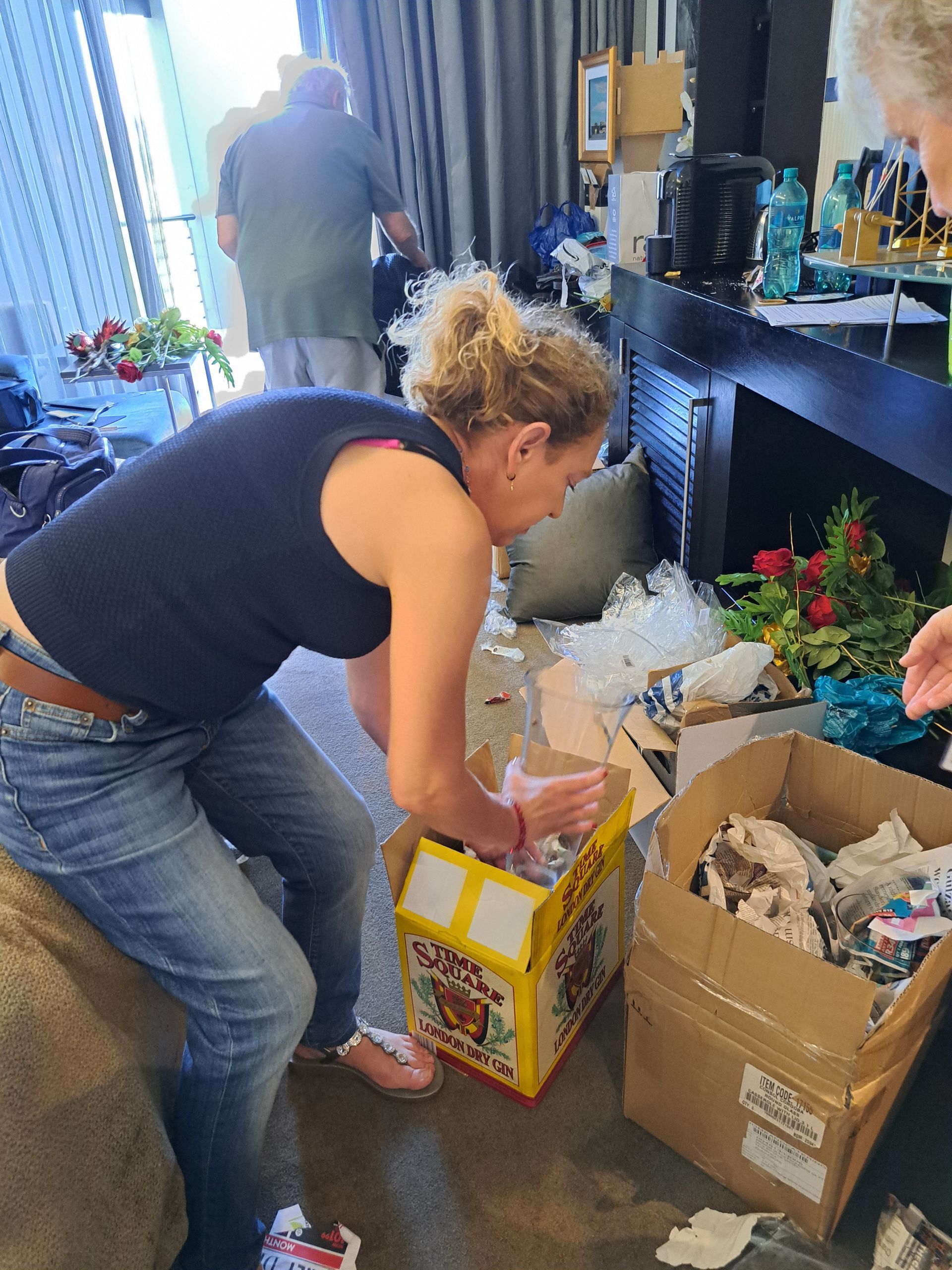 Person packing items into boxes in a cluttered room with plants and a fireplace.