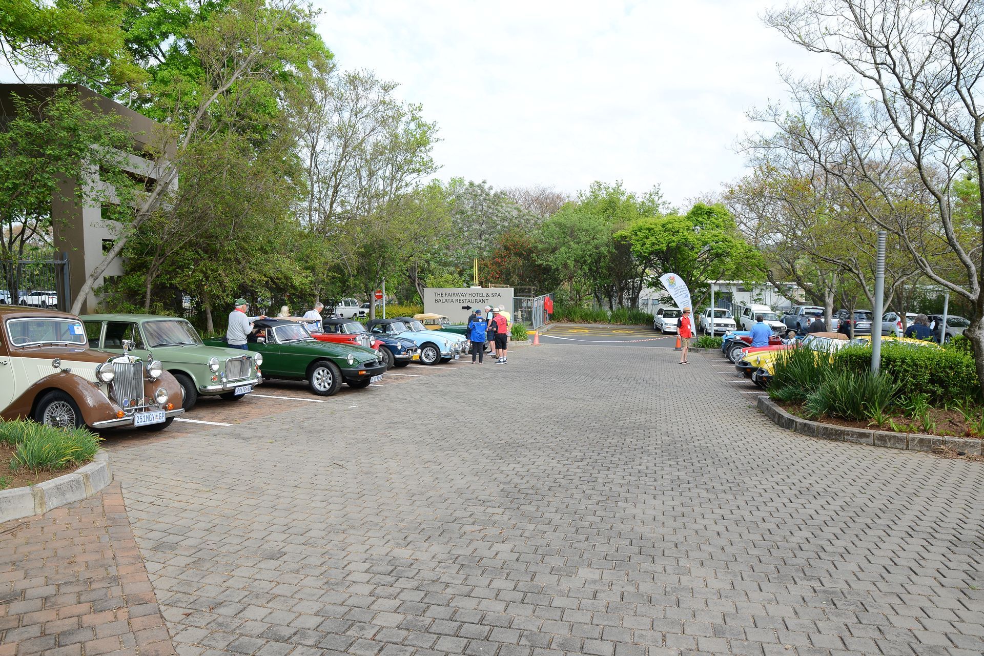 Cars parked along a cobblestone driveway beside trees and buildings in a quiet courtyard.