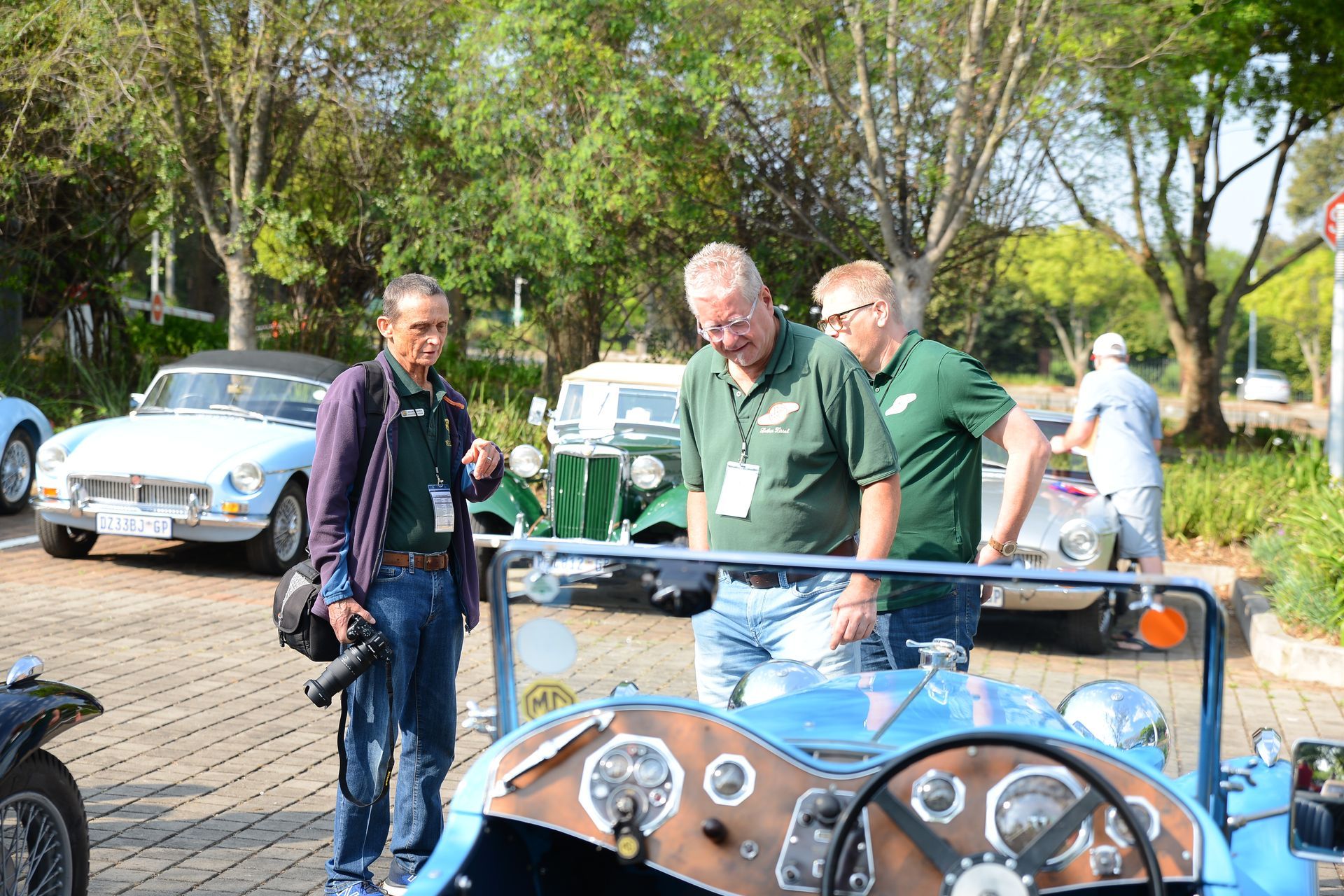 People standing by a vintage car at an outdoor event in a park-like setting