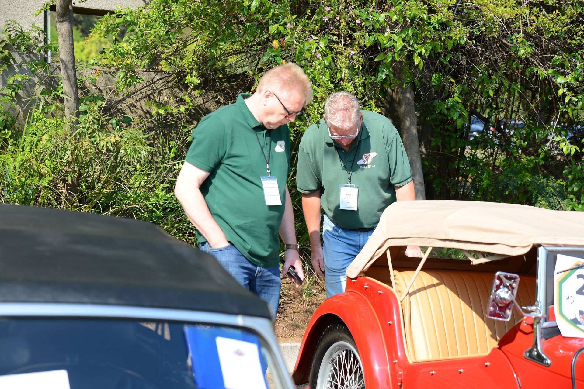 Two people in green shirts look at a red vintage car outdoors.