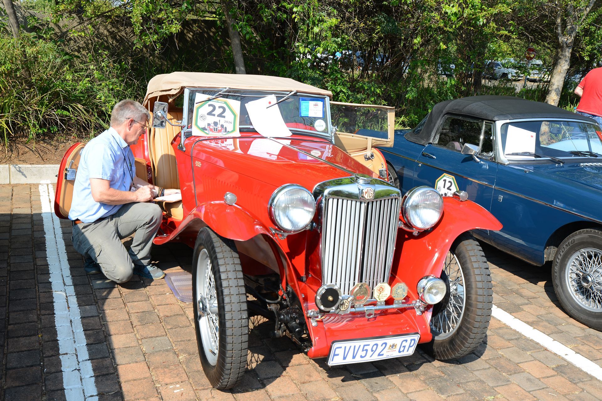 Man kneels beside a red vintage car at an outdoor car show, with a blue car parked nearby.