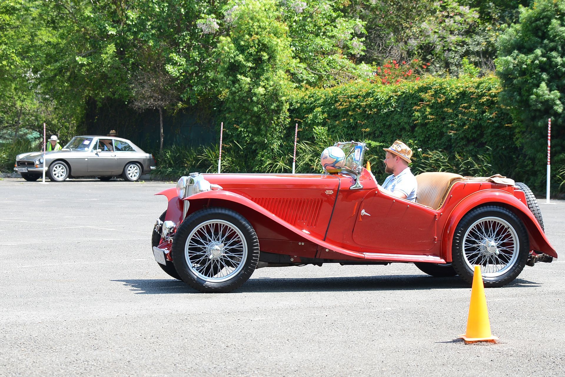 Red vintage convertible with two people driving in a parking lot near a yellow cone.