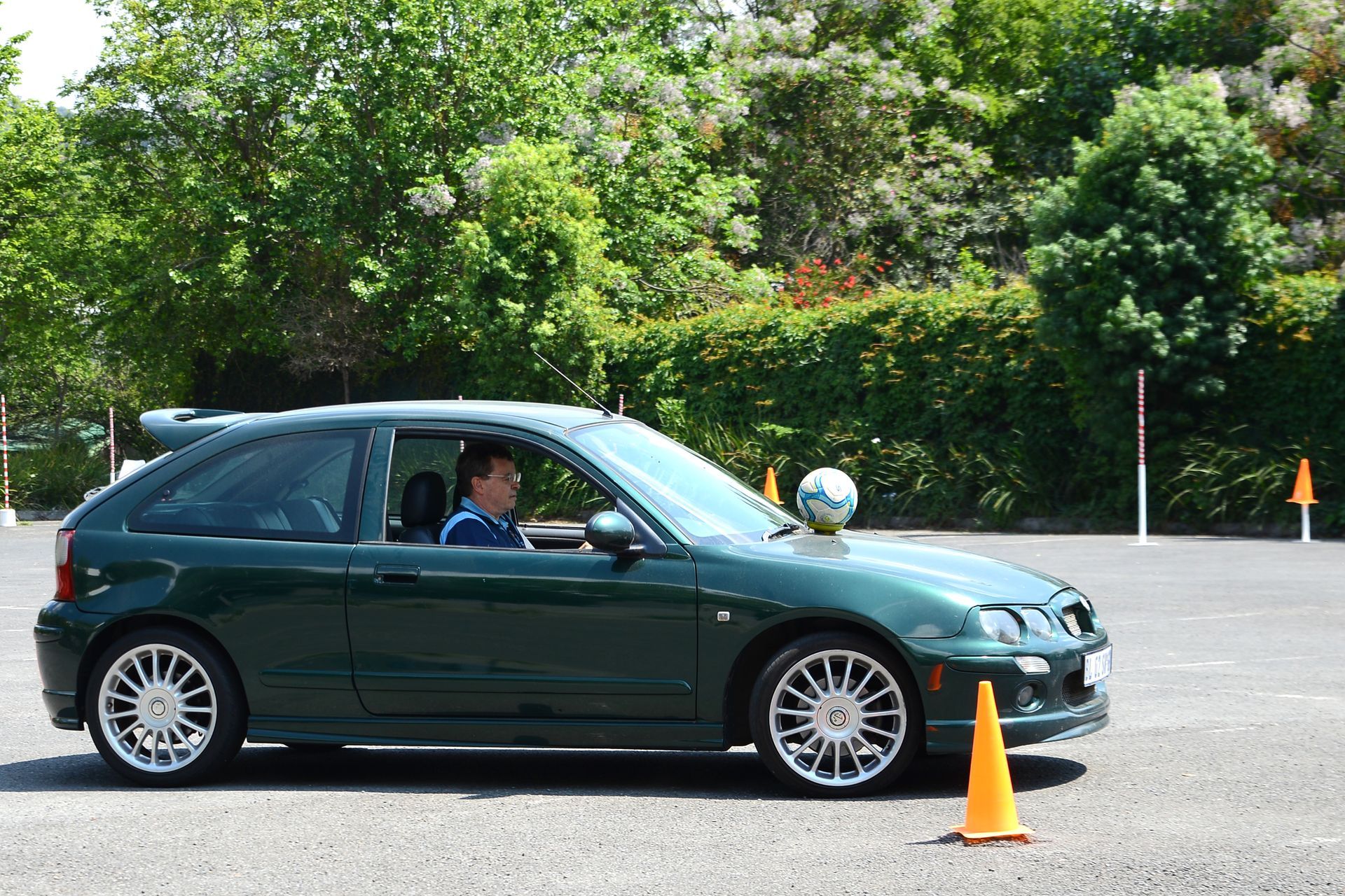 Green sedan with driver stopped beside an orange cone in a parking lot with trees behind it