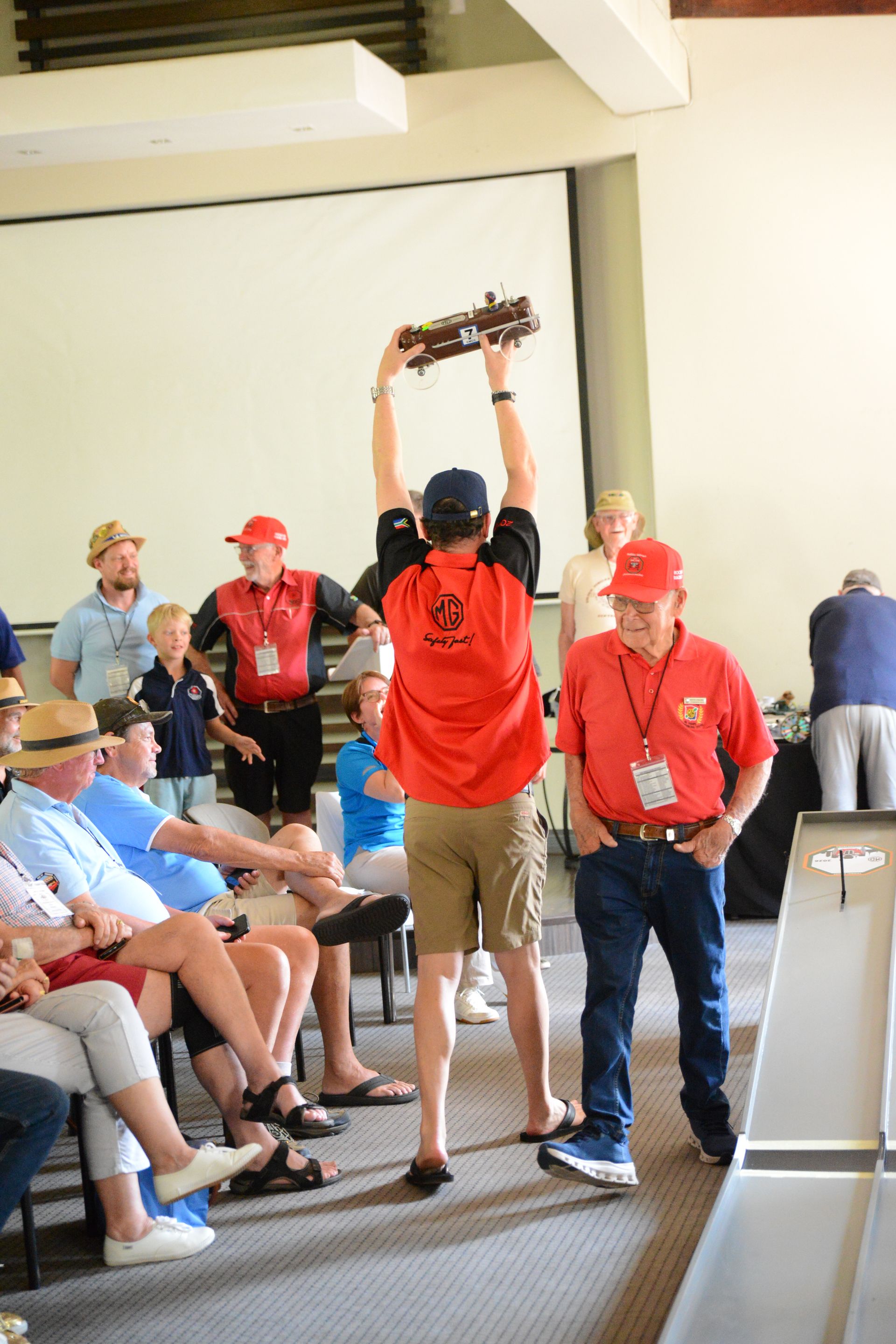 Man in red shirt raises a trophy on a stage while others watch in an indoor hall