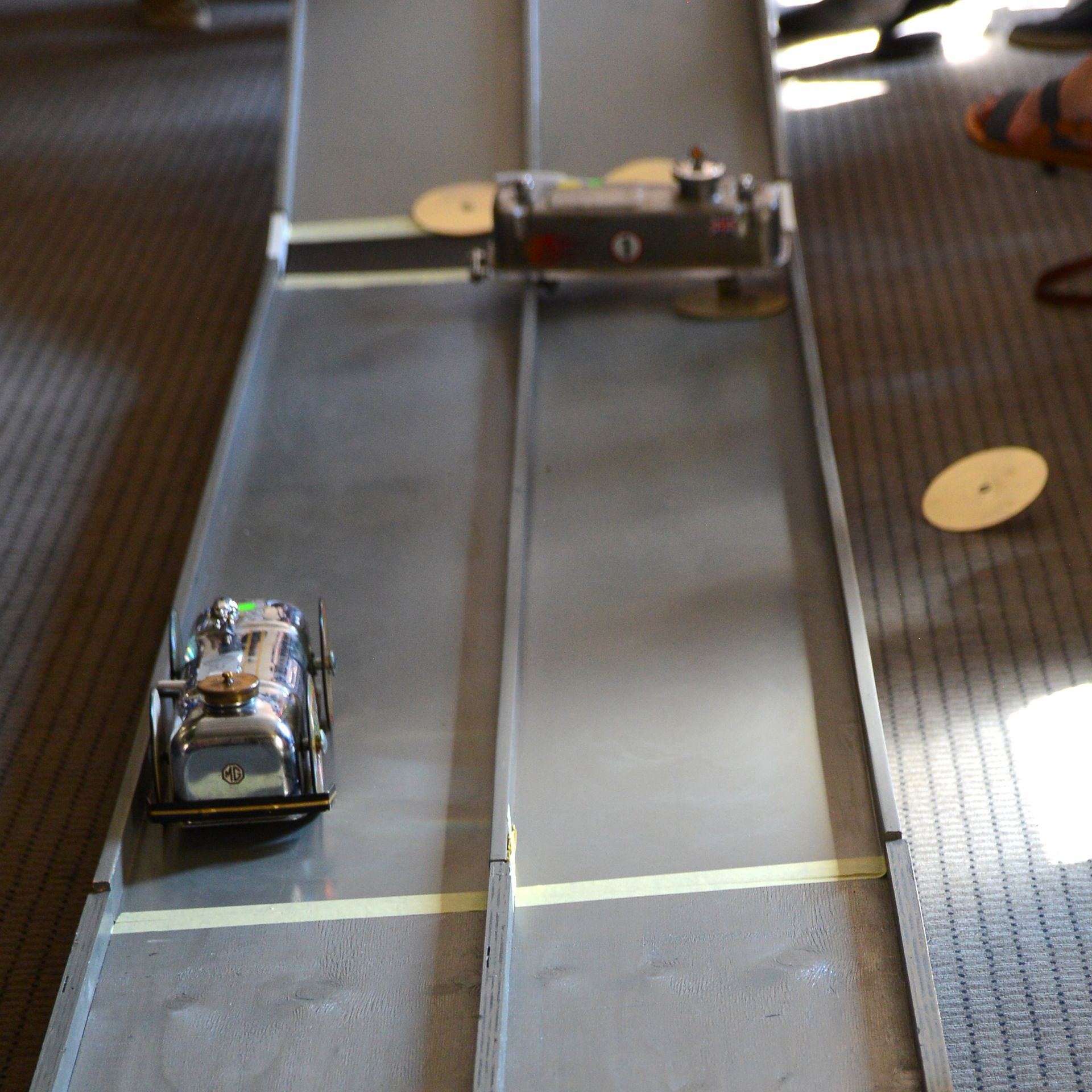 Two toy cars racing on a gray track, one near the bottom left and one ahead at the top center.