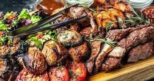 Assorted grilled meats, vegetables, and salad on a wooden platter with tongs and dipping sauces.