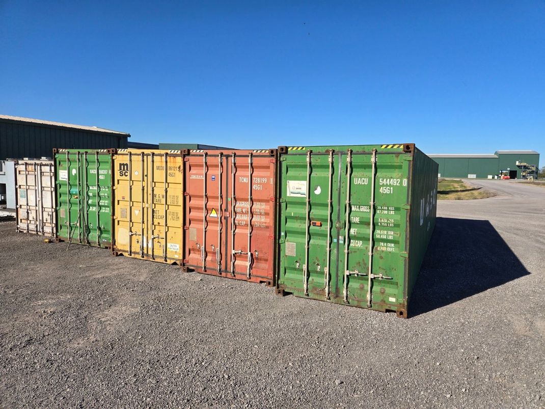 Row of colorful shipping containers on gravel under a blue sky.