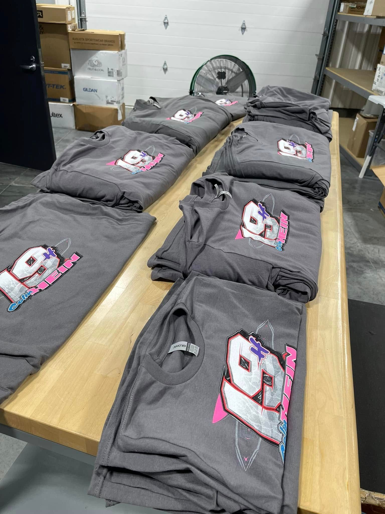 Several gray t-shirts featuring the number 91 printed in red, white, and blue are stacked on a wooden table in a warehouse.
