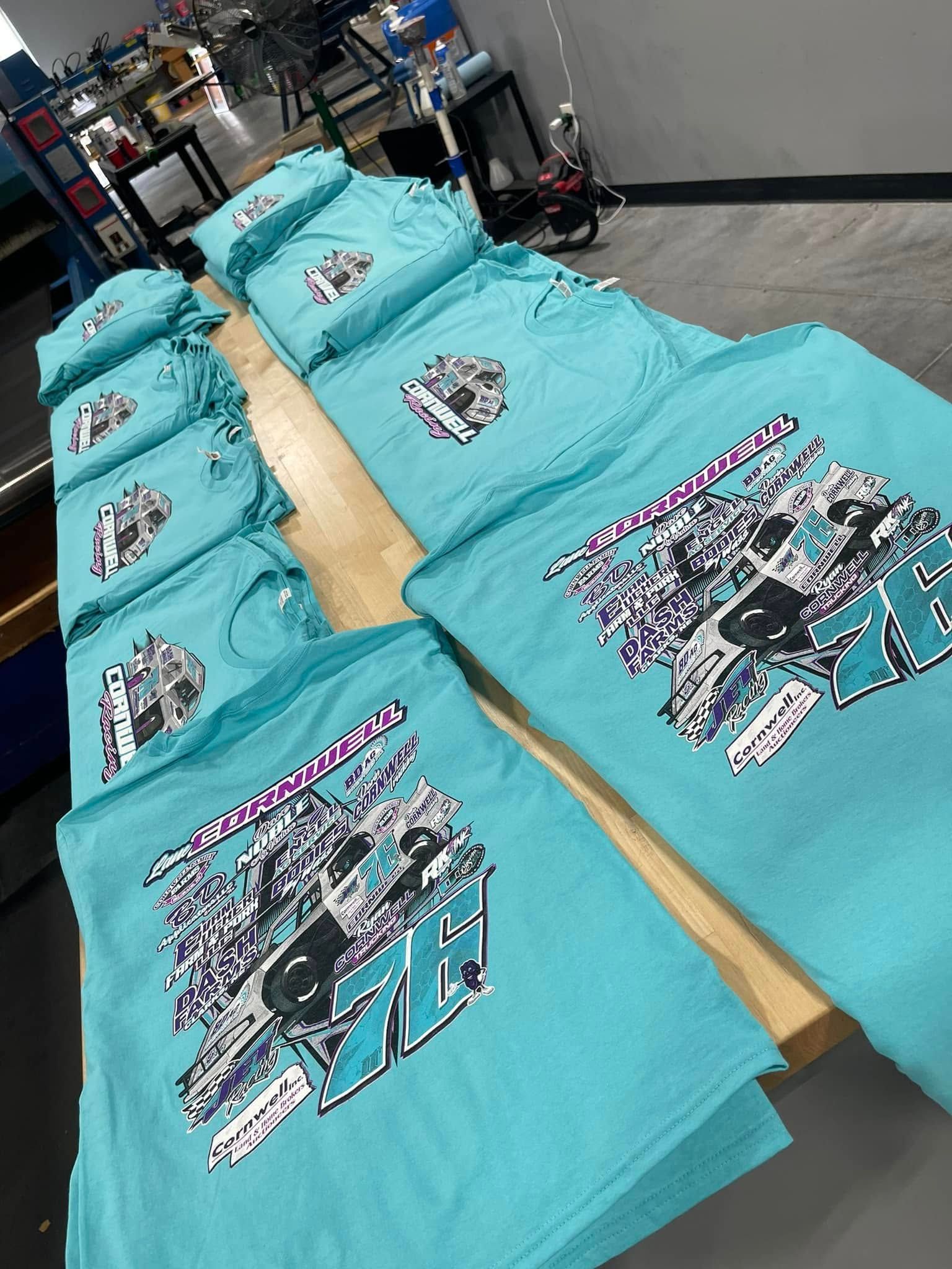 A stack of light blue t-shirts on a wooden table, each featuring a large graphic with a race car and the number 78.