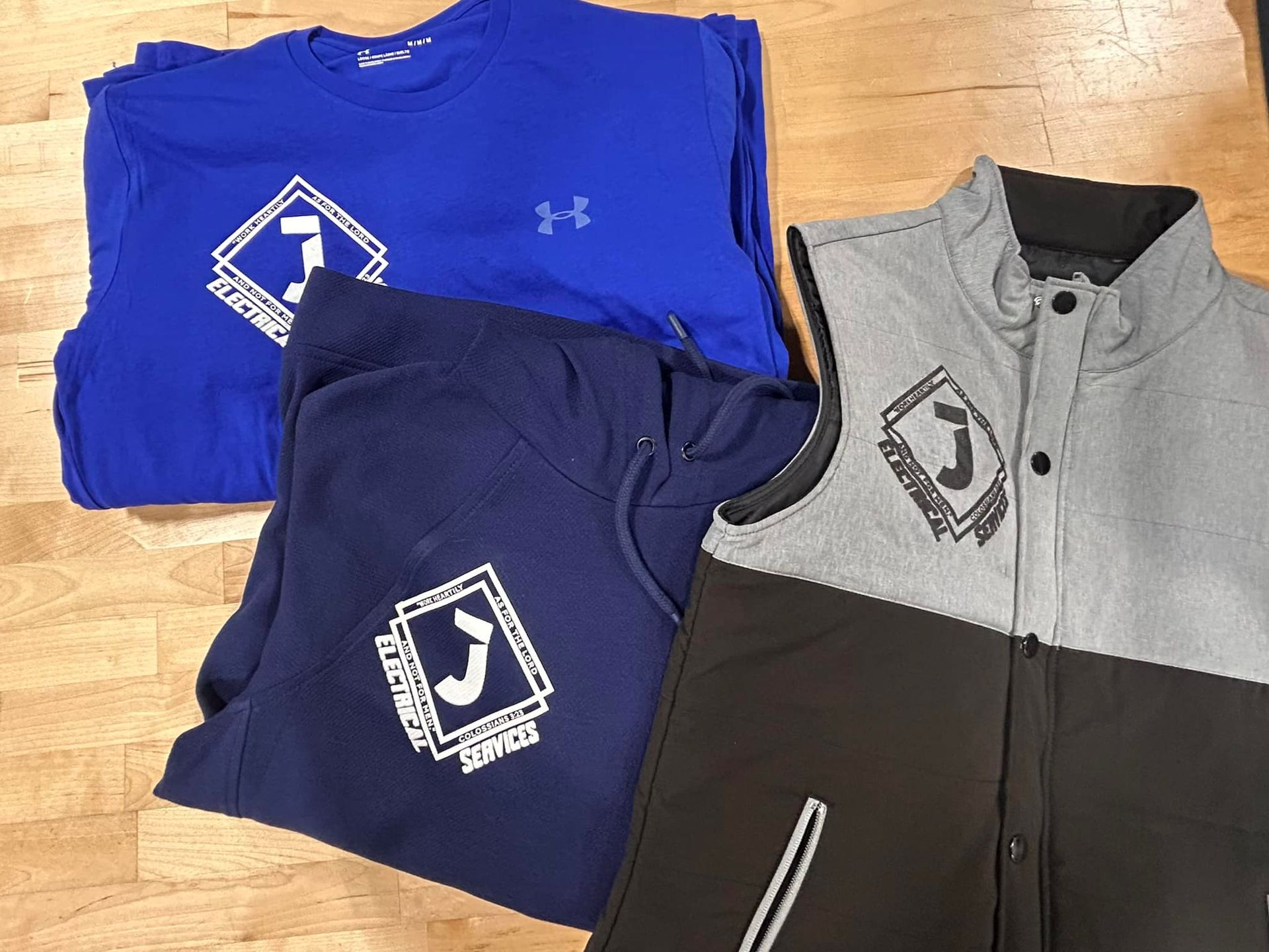 Three articles of clothing with matching logos: a blue Under Armour shirt, a navy hoodie, and a grey and black vest.