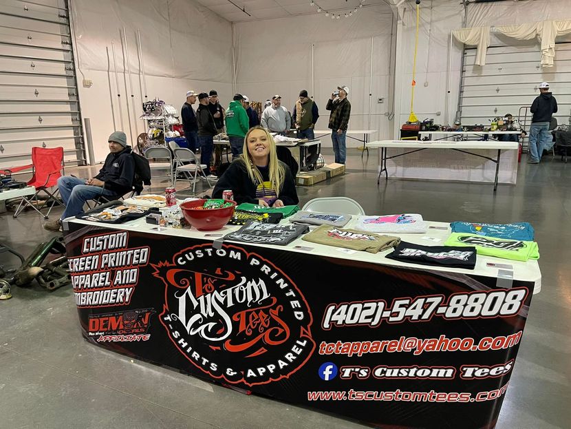 A smiling woman stands behind a custom apparel booth at an indoor event, with merchandise tables and people in the background