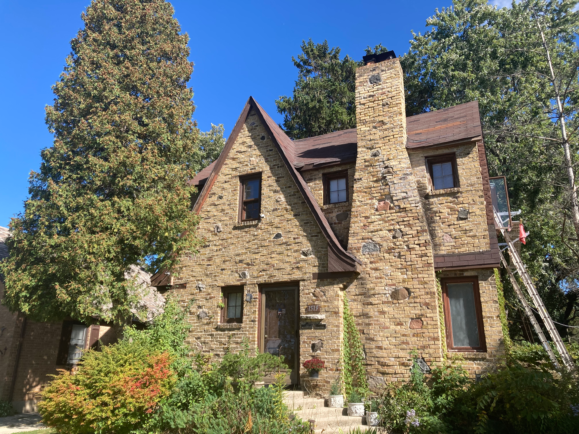 Stone cottage with chimney and steep roof surrounded by trees and greenery under a bright sky