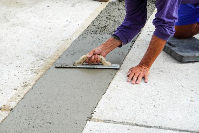 Person using a trowel to smooth wet concrete in a narrow trench on a concrete surface; outdoors.