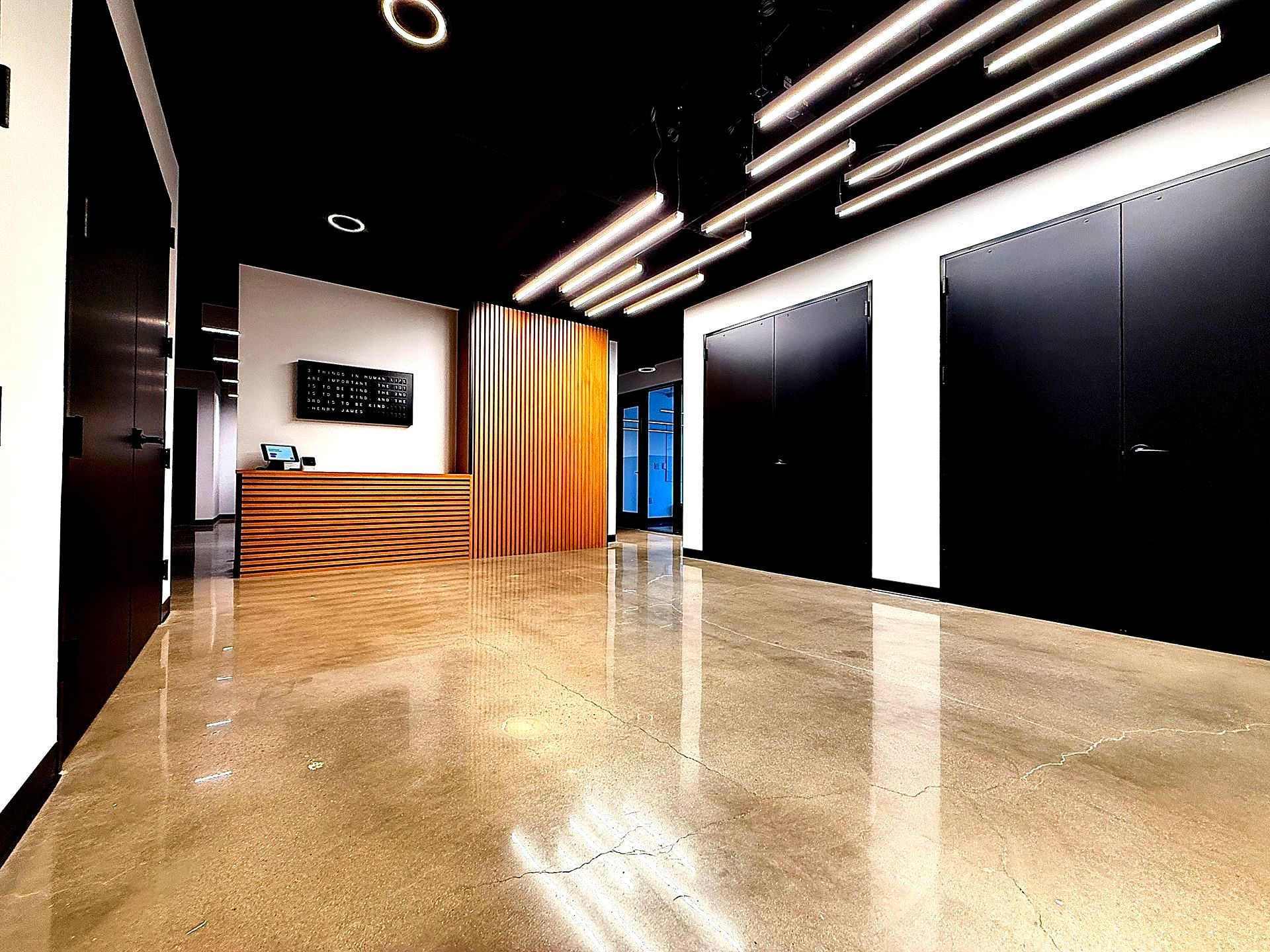 Modern office lobby with polished concrete floor, black doors, and orange/yellow reception desk. Overhead, linear lights illuminate the space.