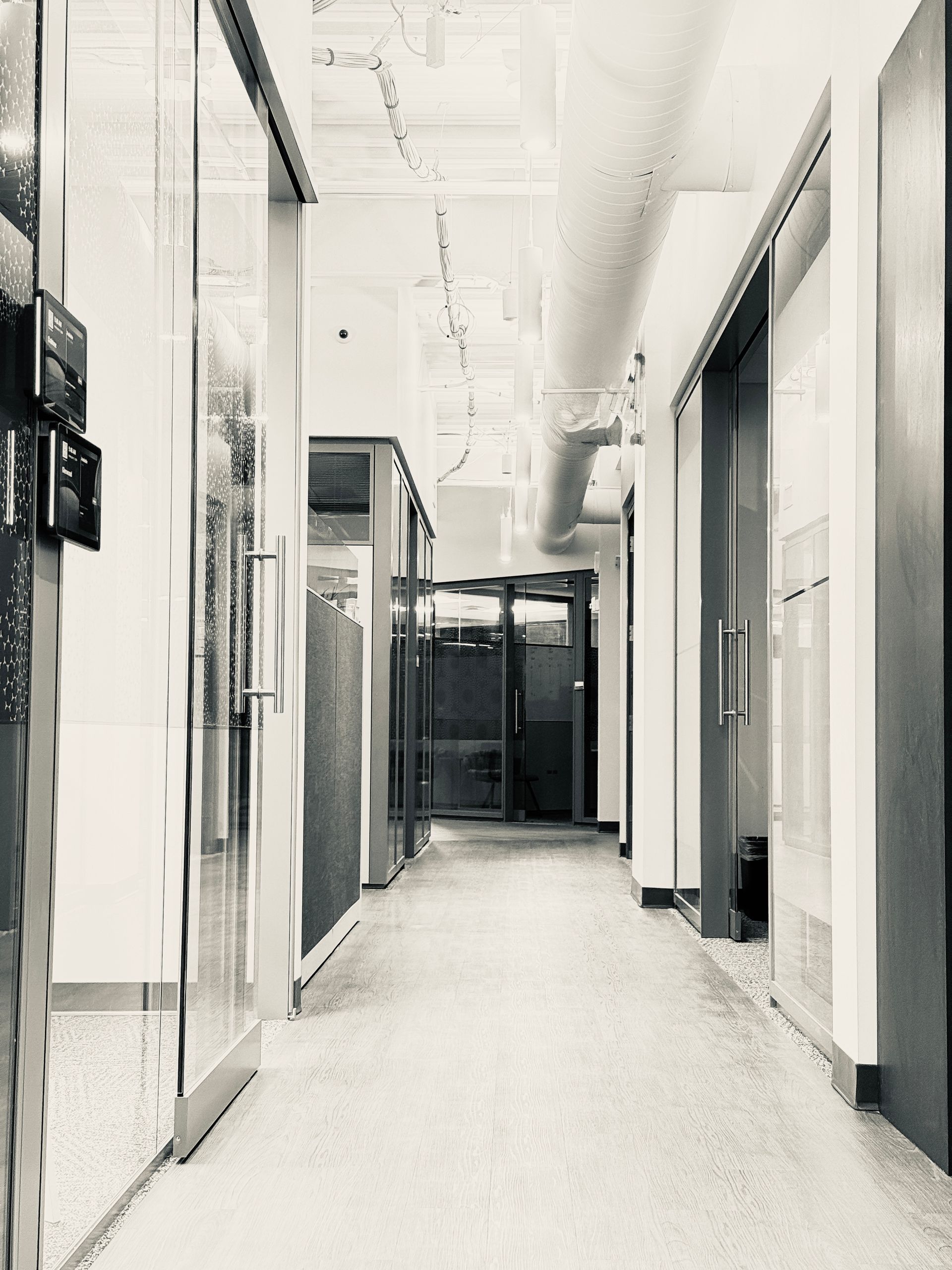 A long, empty office corridor with glass-walled rooms on either side and a distant doorway, all in grayscale.