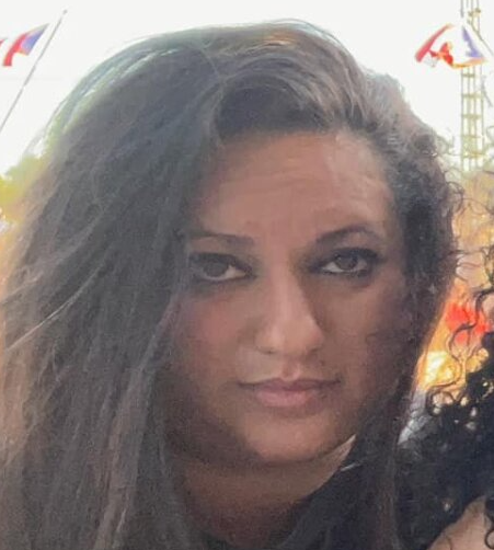 Woman with long dark hair, looking at the camera. Outdoor setting with flags and structures in the background.