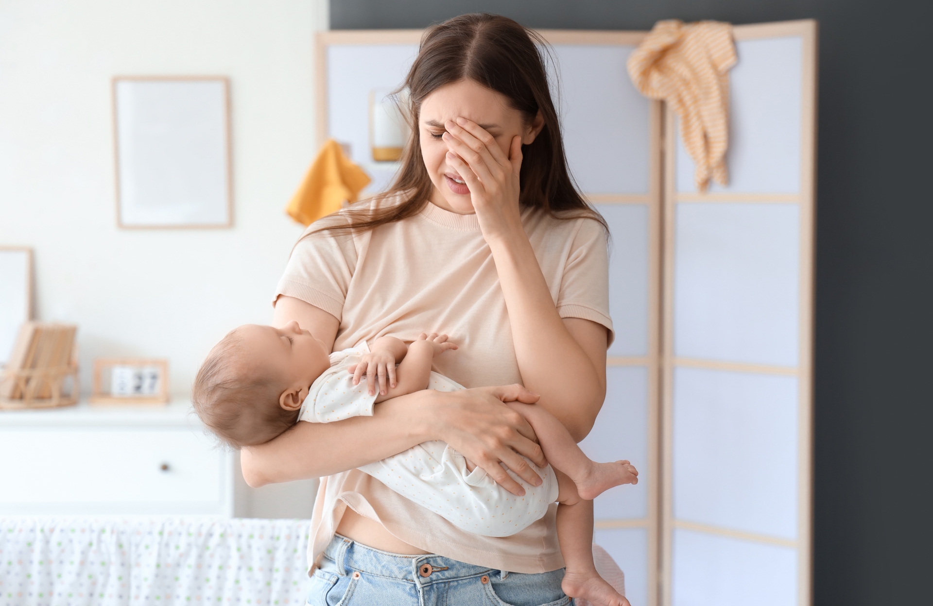 A person holding a sleeping baby in a bright room, with their hand over their face looking distressed.