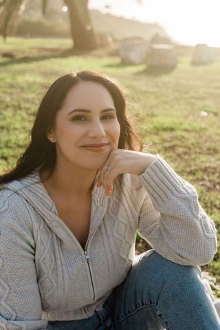 A person in a light grey cable-knit sweater sits in a grassy outdoor area, resting their chin on their hand and smiling.