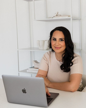 A person sitting at a desk with an open laptop, smiling in front of a white shelf with decorative items.