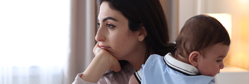 A person resting their chin on their hand with a pensive expression, holding a baby against their shoulder indoors.