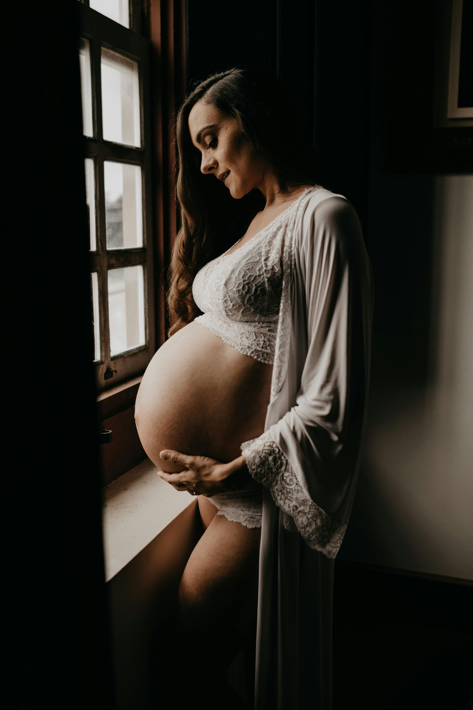 A pregnant person in a white lace robe stands by a window in a dimly lit room, gently holding their abdomen.