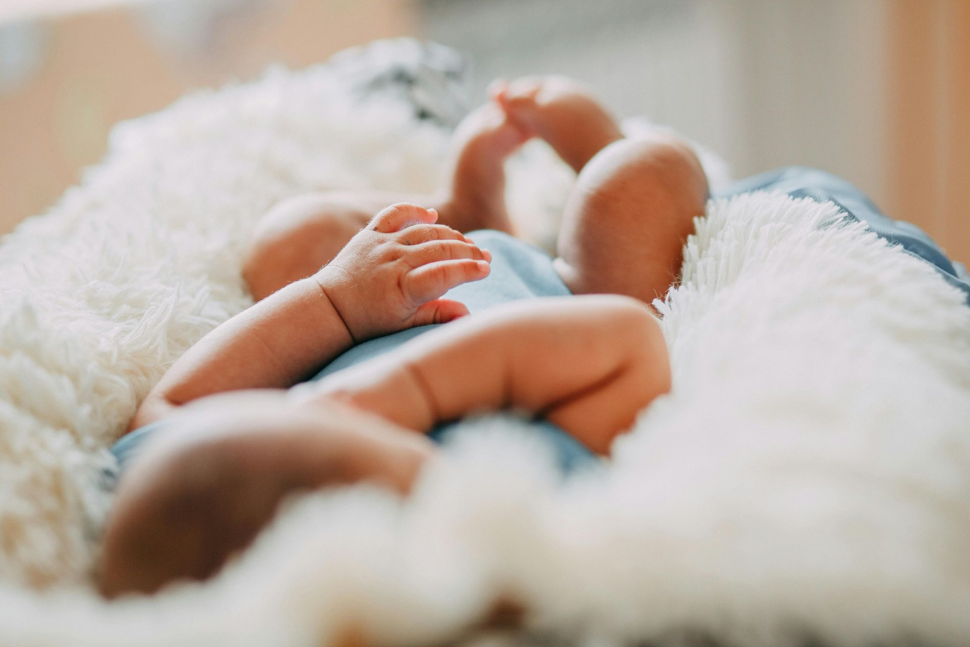 A newborn lying on a soft, fluffy white blanket, wearing a blue outfit with legs and arms curled up.