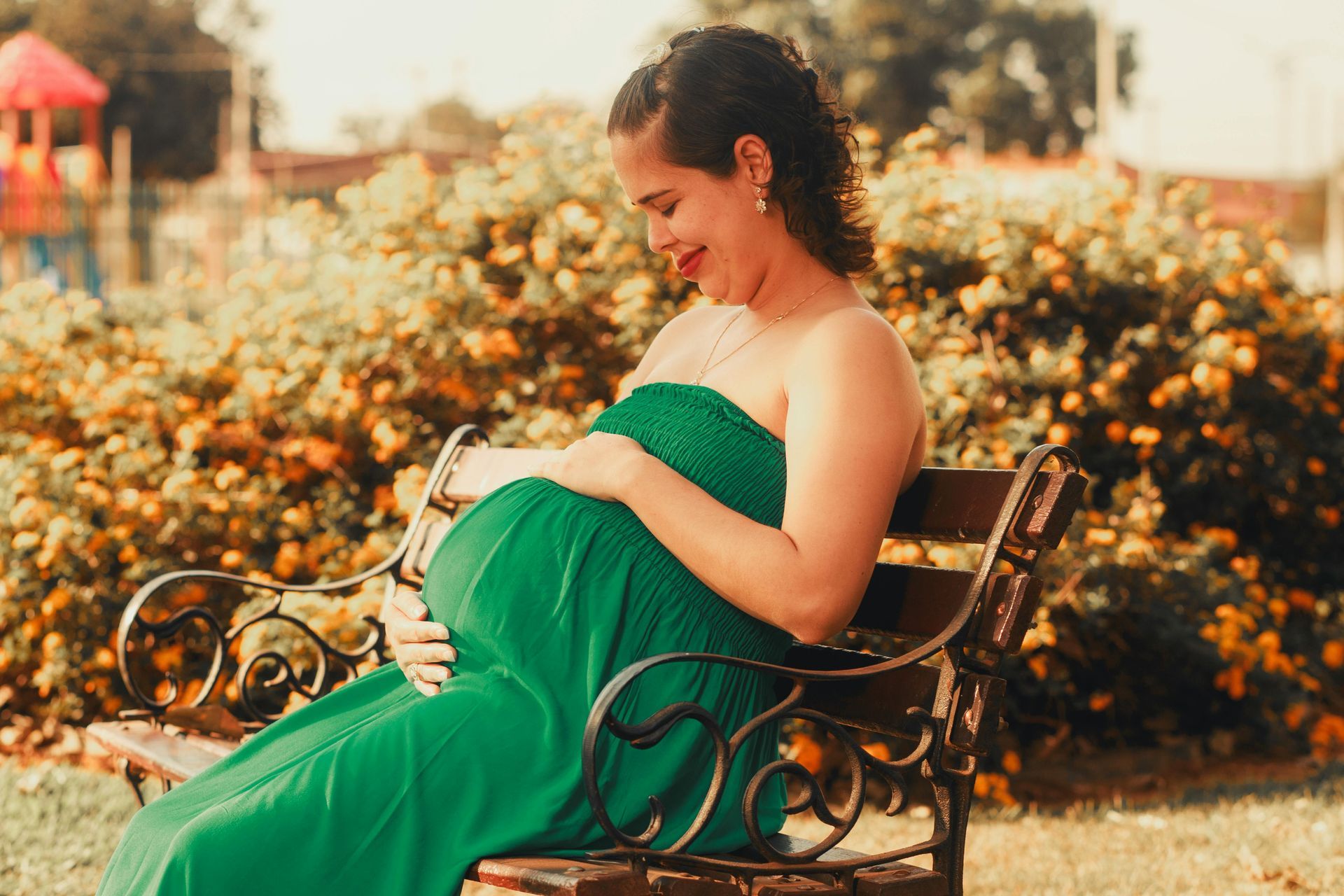 A pregnant person in a bright green strapless dress sitting on a park bench, looking down at their belly.