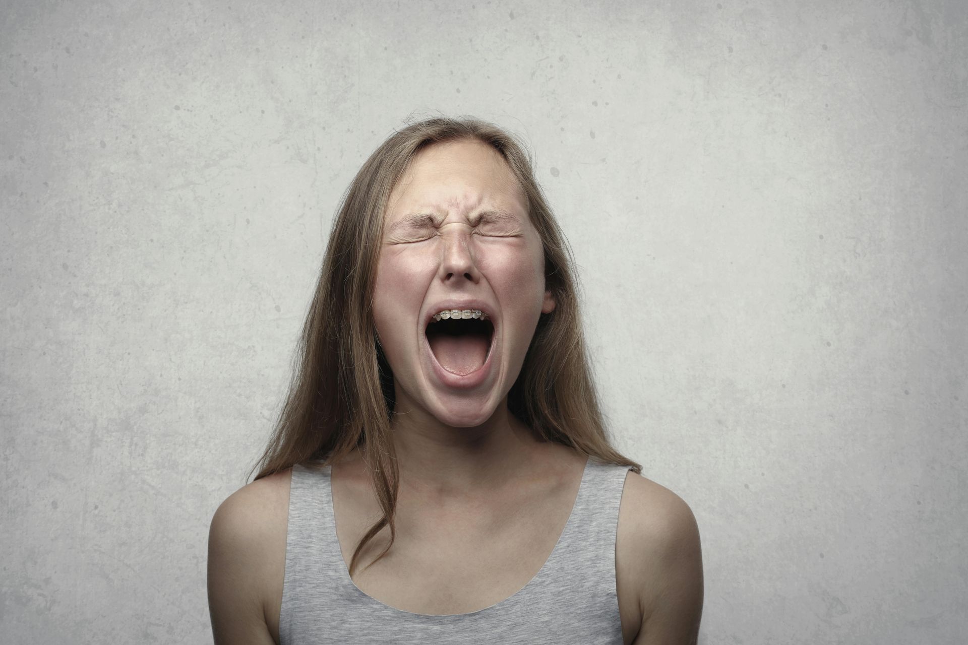 A person with long hair against a light gray background, mouth wide open with eyes squeezed shut in an expression of agony.