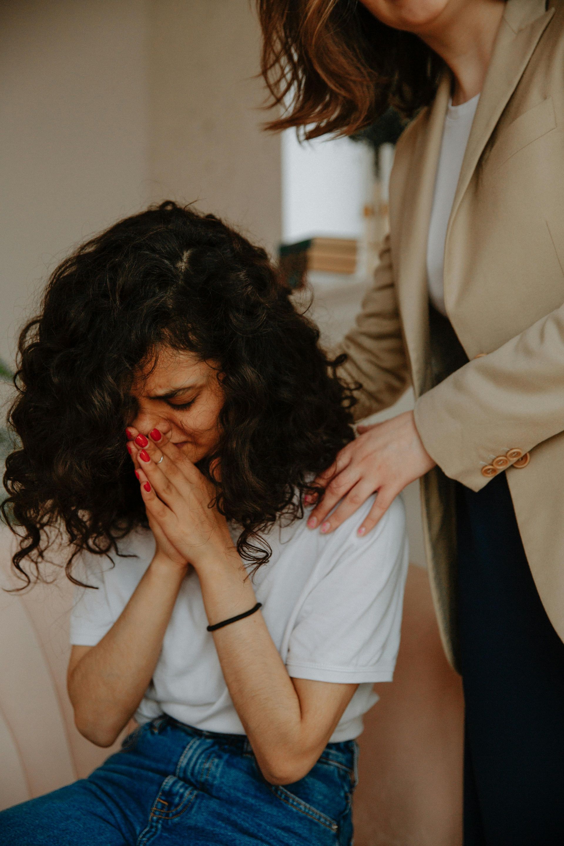 A person with curly hair covers their face in distress while another person comforts them with a hand on their shoulder.