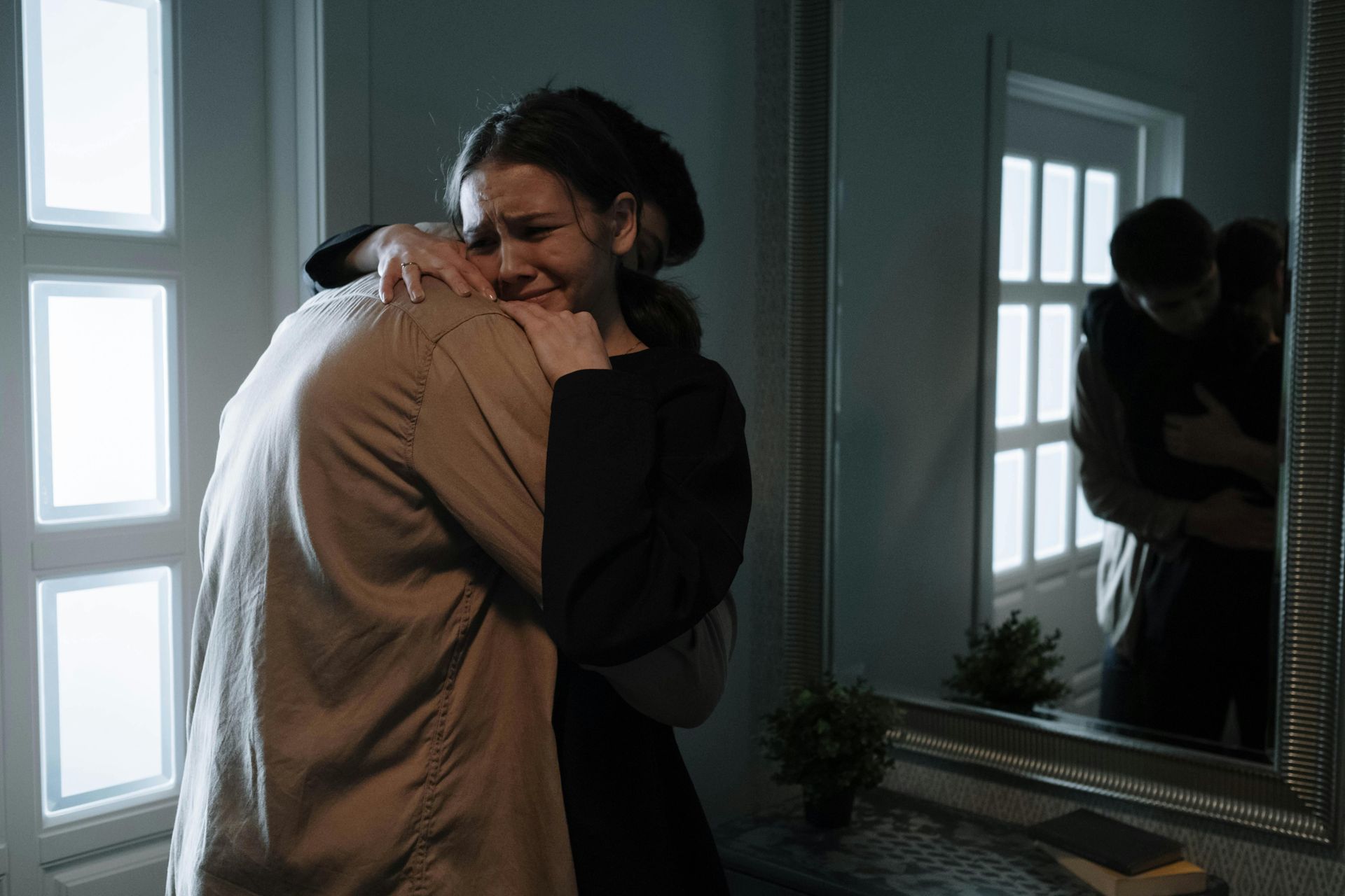 Two people embrace in a dimly lit room, with their reflection visible in a mirror on the wall.